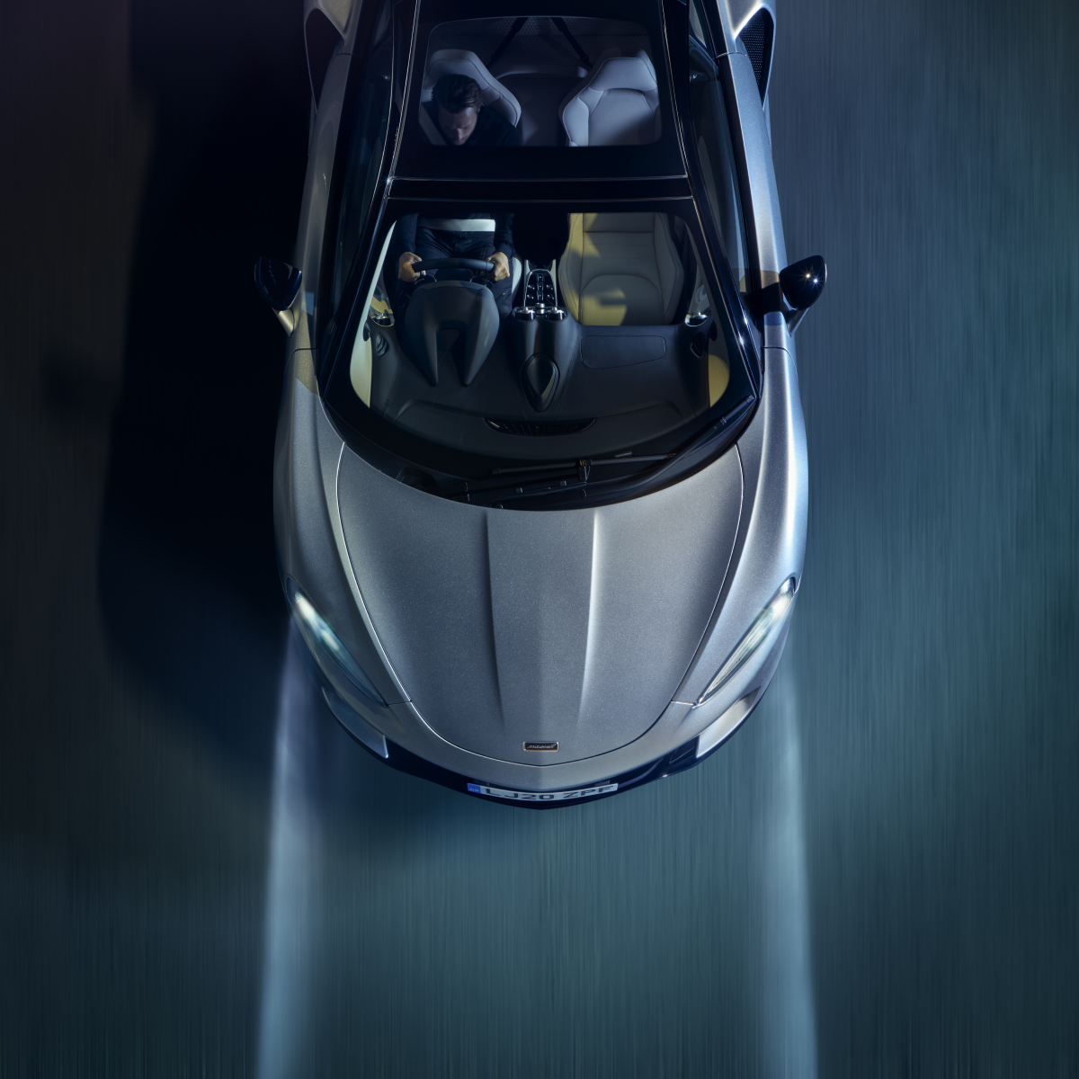 A high-angle view of a silver McLaren GT with its headlights on, driving at night with a motion blur effect on the road.