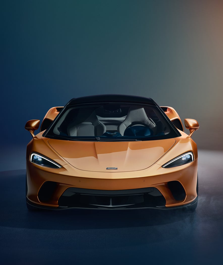 A front-on view of the McLaren GT in a bespoke gold finish, with its headlights on, in a dark studio setting.