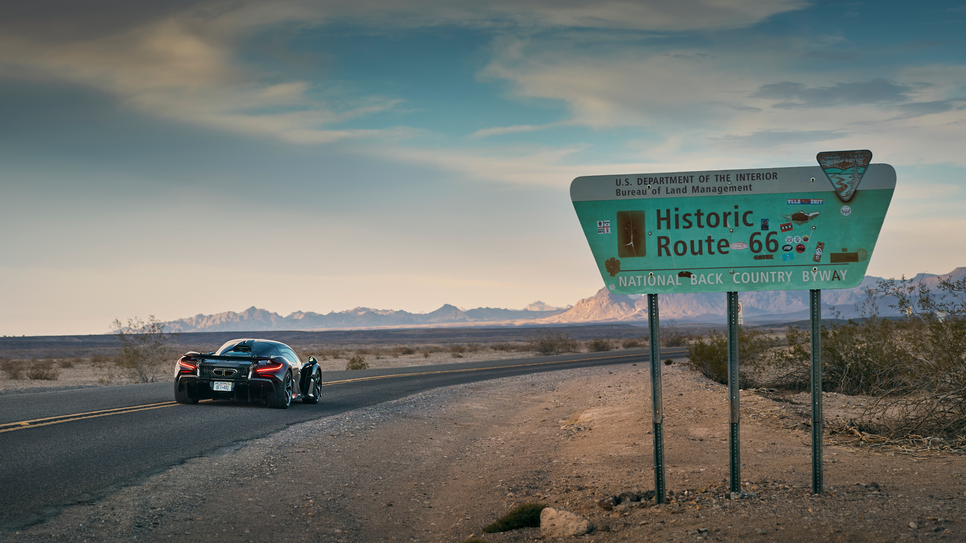 McLaren W1 prototype driving on a road near a Historic Route 66 sign in a vast desert landscape. Rear view of the Ultimate Series car, emphasizing long-distance durability and real-world hot weather testing for its 1275PS hybrid engine.