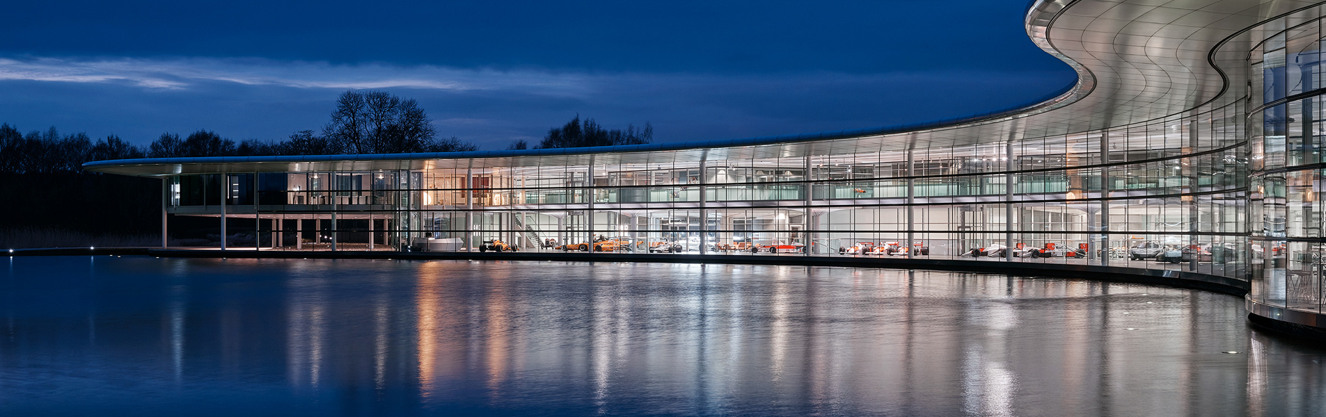 A nighttime photograph of the McLaren Technology Centre reflected in the large lake in front of it.