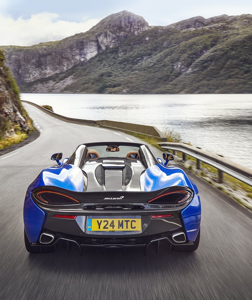 The McLaren 570S Spider, with its top down, driving on a winding road with a large lake and mountains in the background.