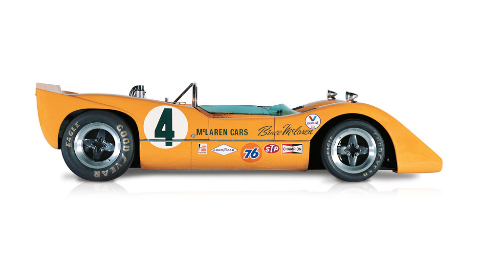 Side view of the orange McLaren M6A, with the number 4 painted on the side of the body and covered in sponsorship stickers, on a white background.