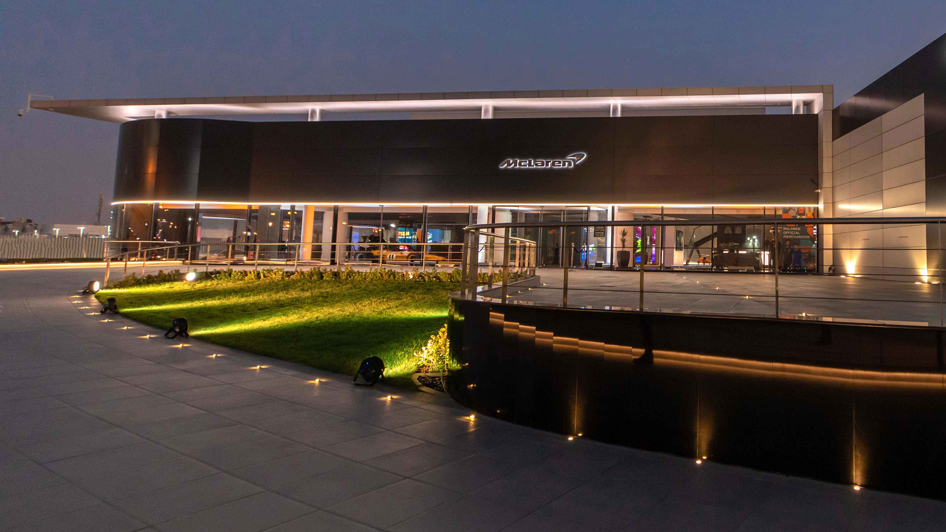 McLaren Bahrain official retailer exterior at night. The modern, dark-clad luxury supercar showroom features illuminated branding and architecture over a large tiled plaza.