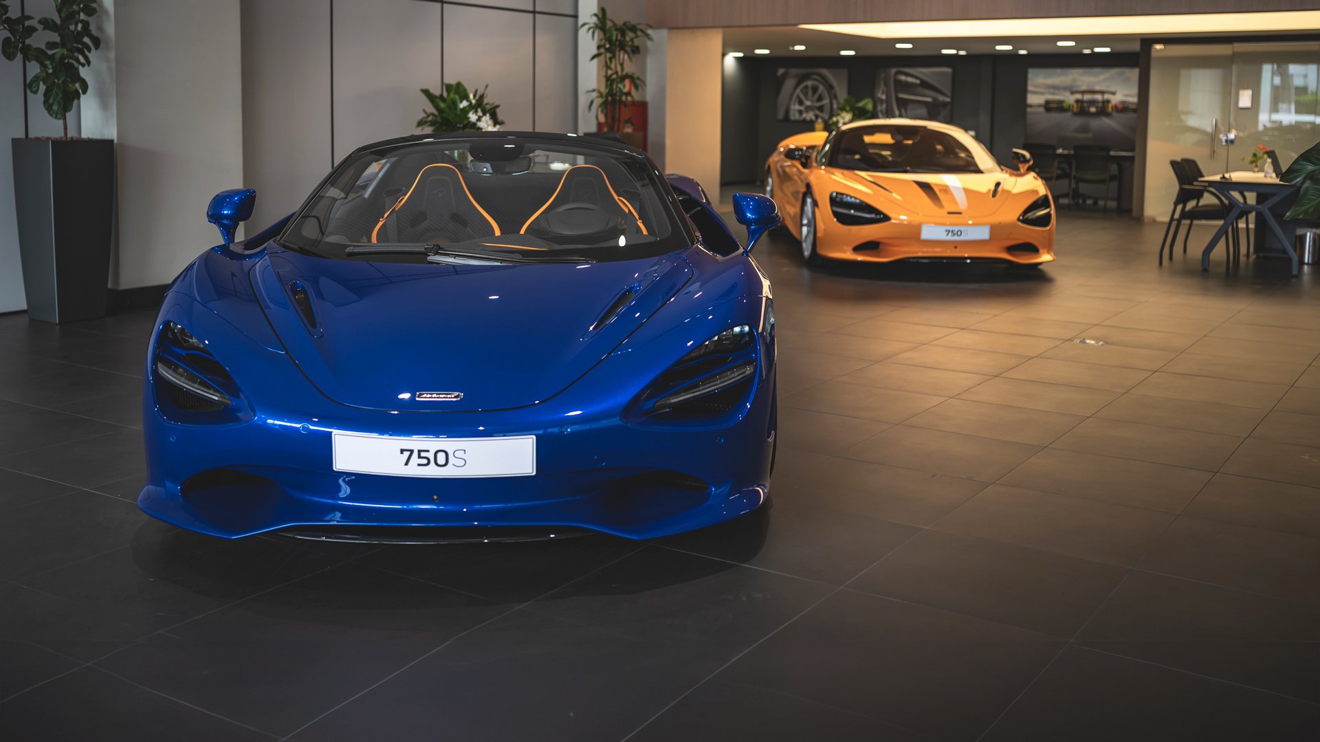 McLaren 750S Spider convertible in striking Aurora Blue (foreground) and a McLaren 750S Coupe in signature McLaren Orange (background), displayed side-by-side in a modern supercar showroom.