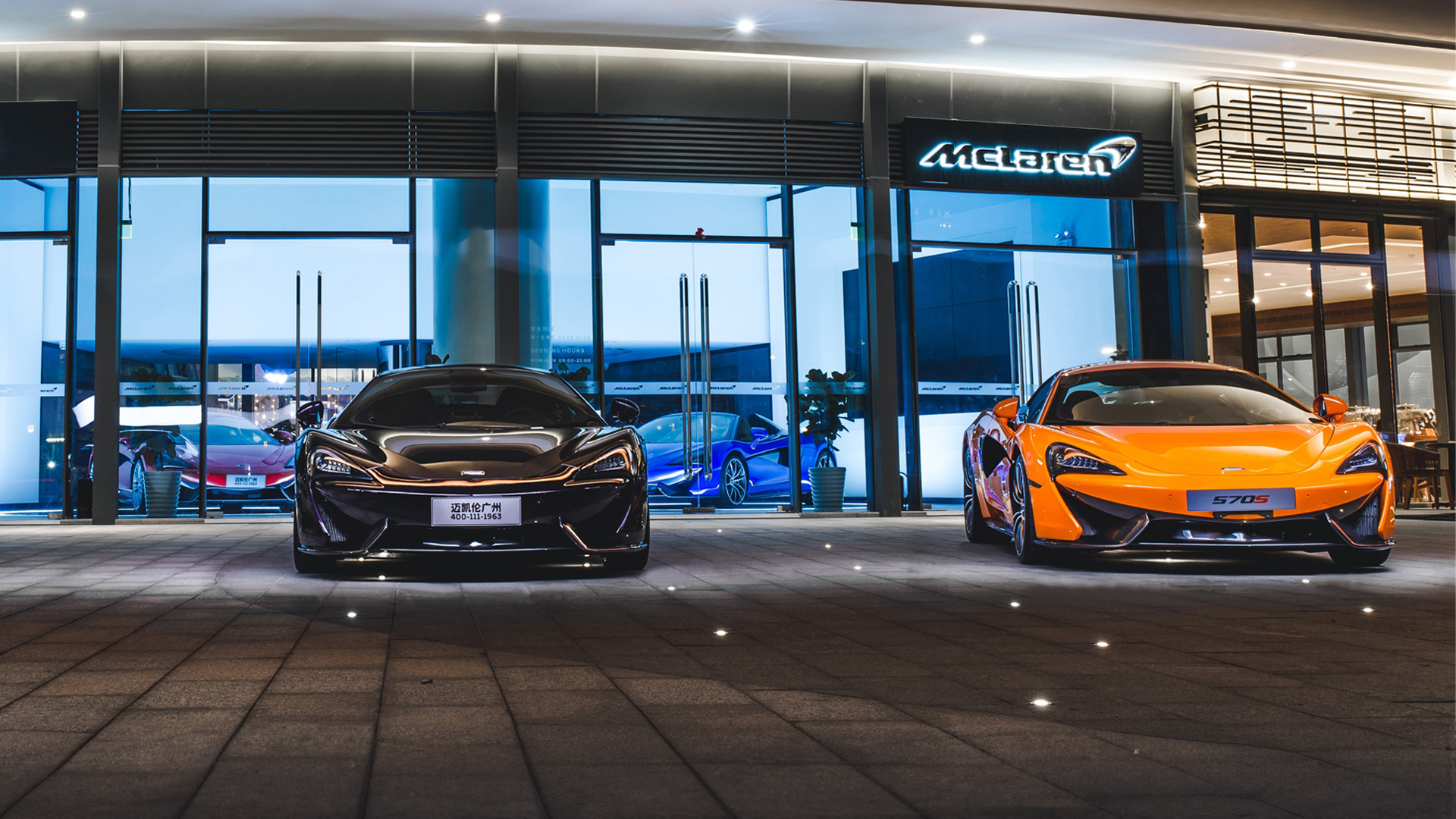 McLaren Guangzhou, China retailer exterior at night, showcasing two supercars. A dark metallic grey McLaren (left) and a bright McLaren Orange 570S (right) are parked outside the modern, glass-fronted showroom.