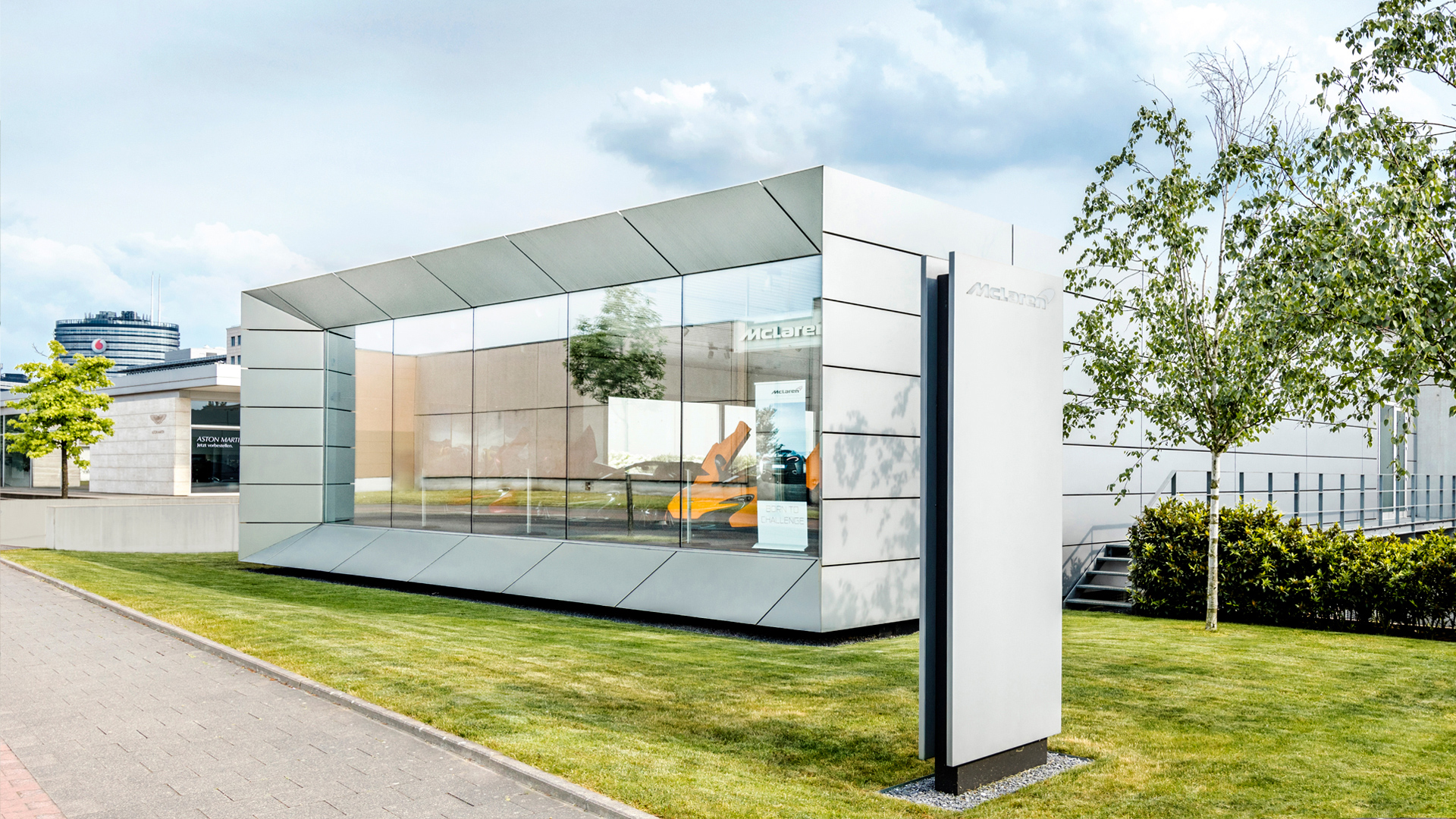 McLaren Düsseldorf (Moll Sportwagen) official retailer exterior. The showroom features sleek, modern glass and silver panel architecture with an orange McLaren Artura supercar visible through the large window.