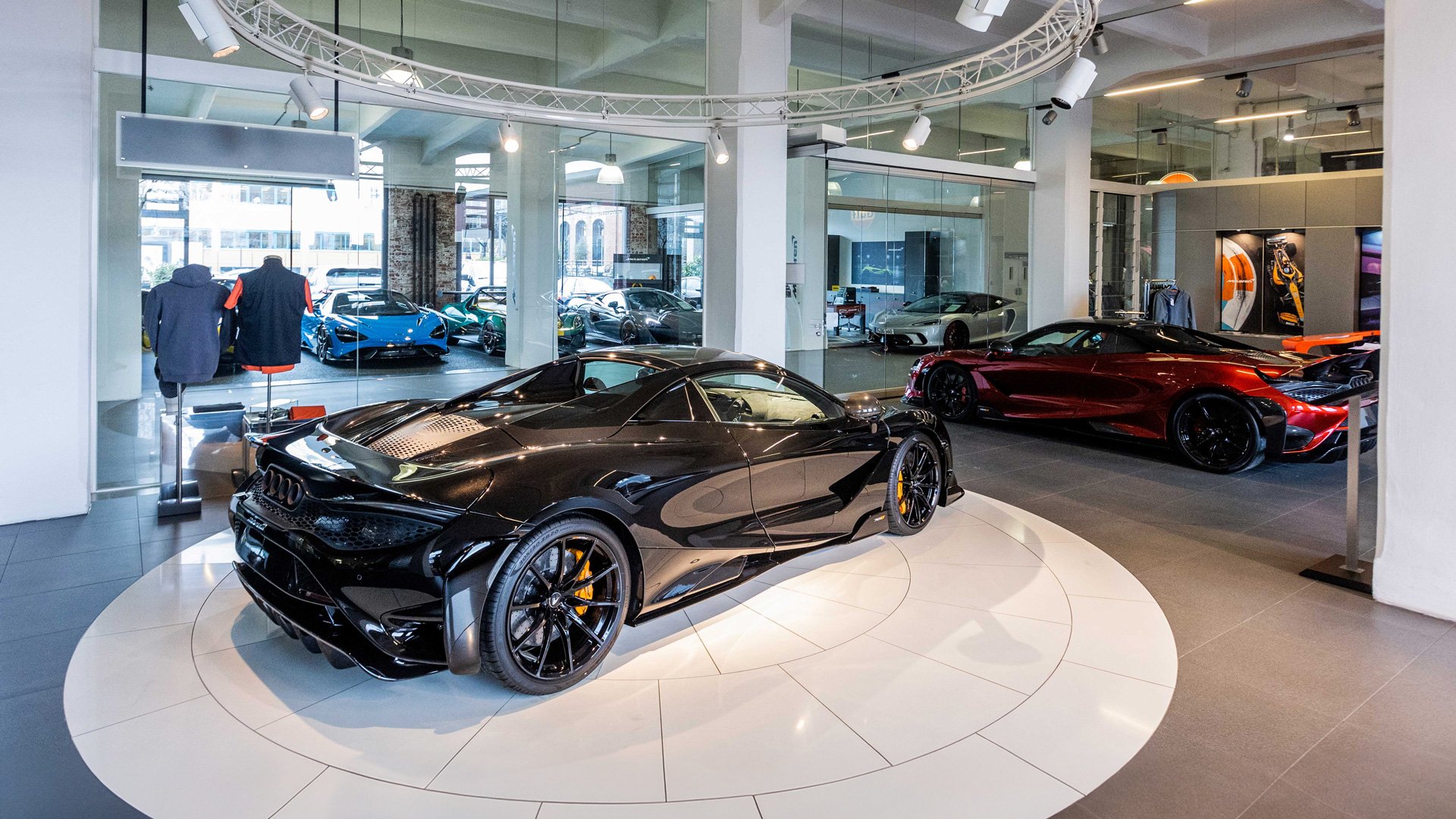 McLaren Frankfurt official retailer showroom interior. A black McLaren 765LT supercar with yellow brake calipers is displayed prominently on a rotating platform next to a metallic red McLaren.