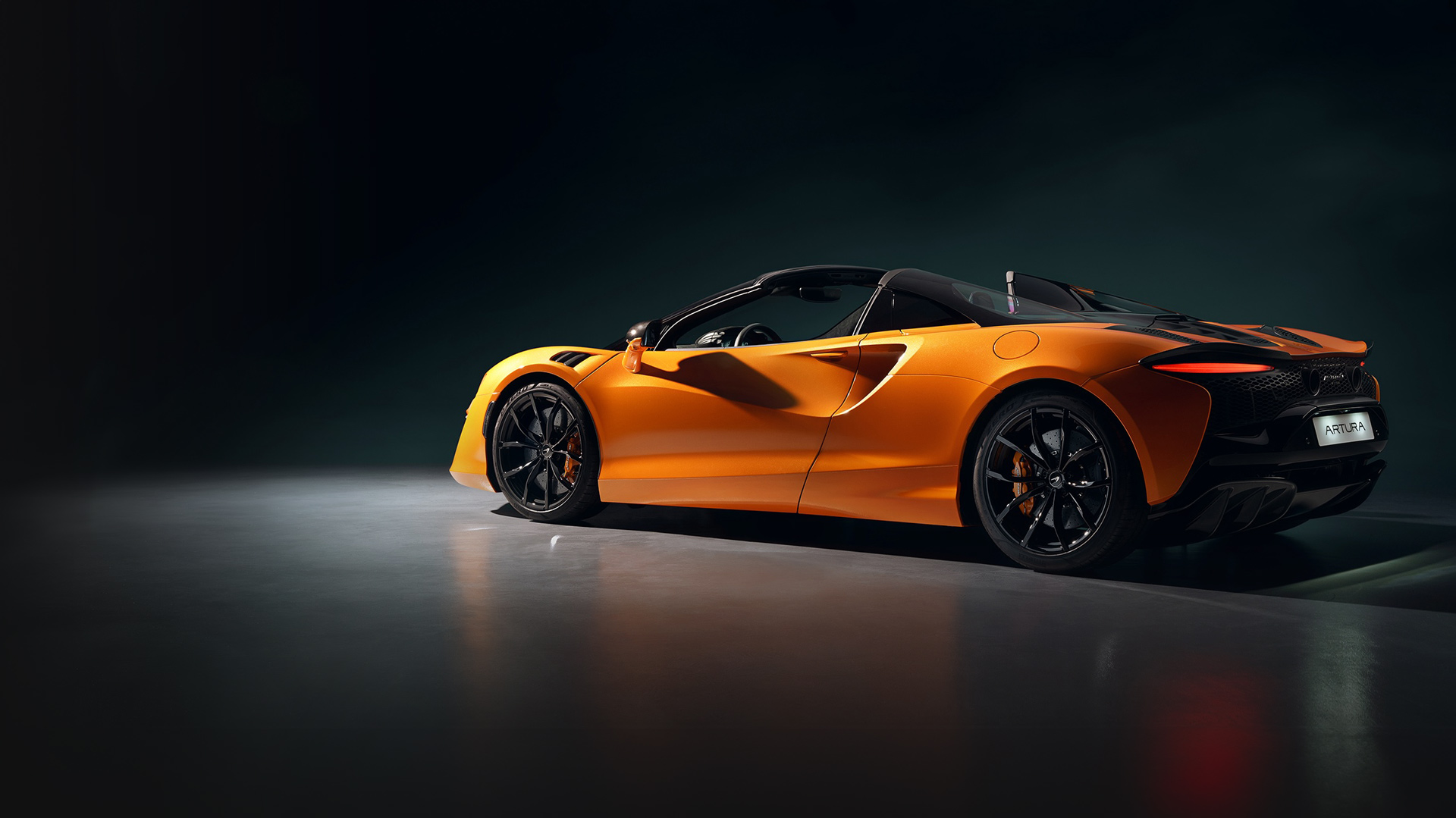McLaren Artura Spider High-Performance Hybrid convertible in a vibrant McLaren Orange or Papaya Spark paint, displayed in a dimly lit studio or showroom.
