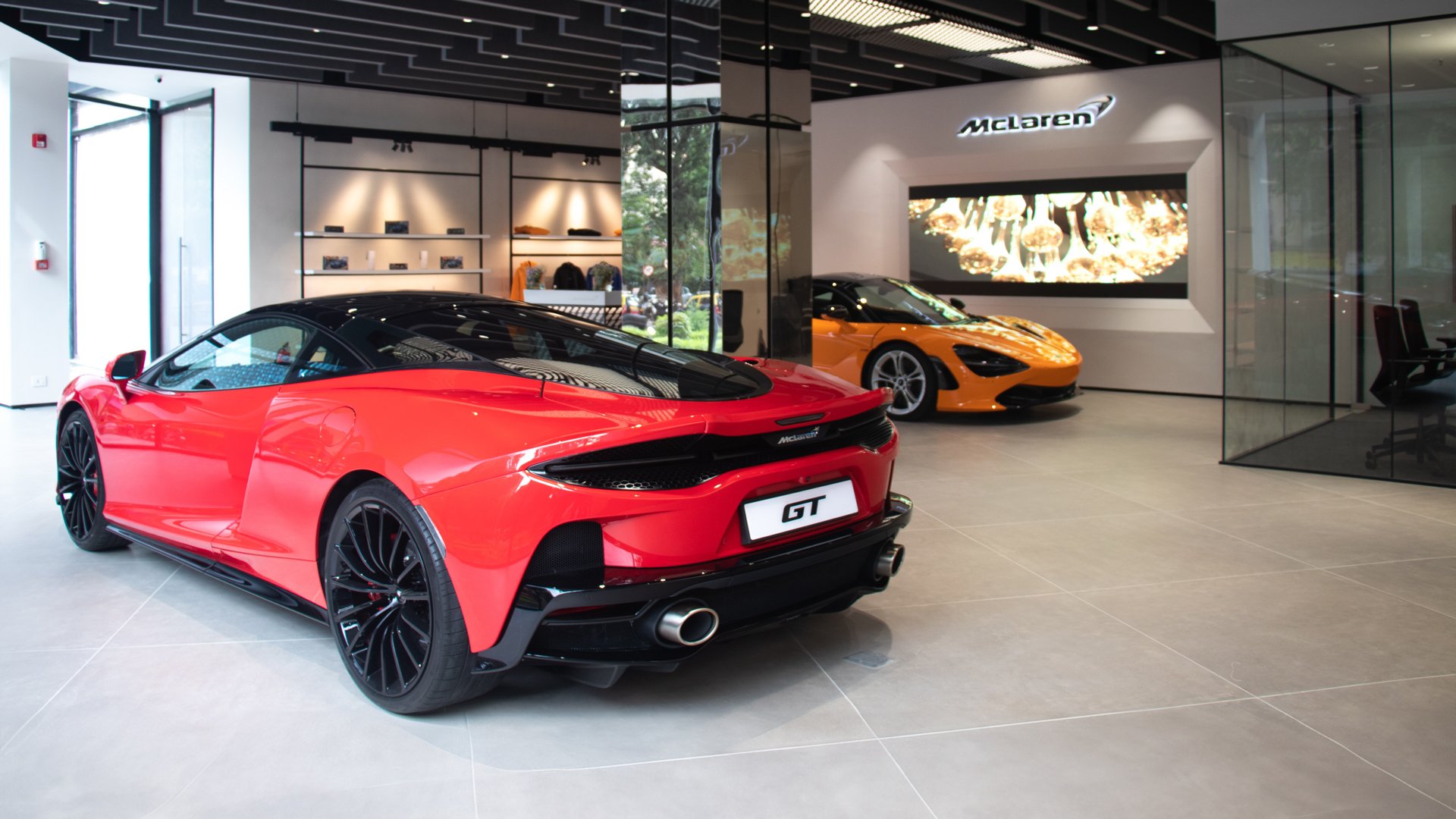 McLaren Mumbai official retailer showroom interior. A red McLaren GT Grand Tourer is parked in the foreground, with an orange McLaren 750S supercar in the background.