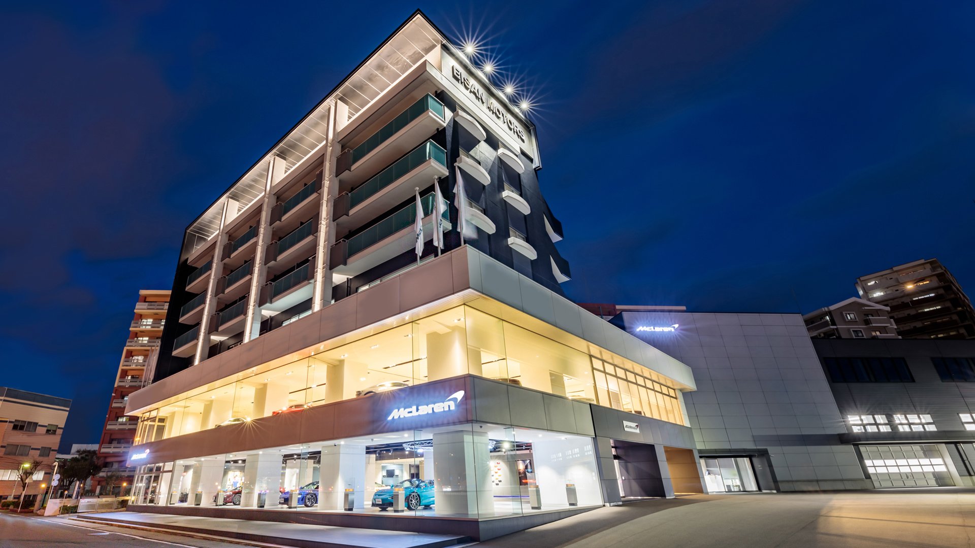 McLaren Fukuoka official retailer exterior at night in Japan. The modern, multi-story glass and concrete building features the McLaren logo prominently, with luxury supercars visible on the brightly lit ground-floor showroom.