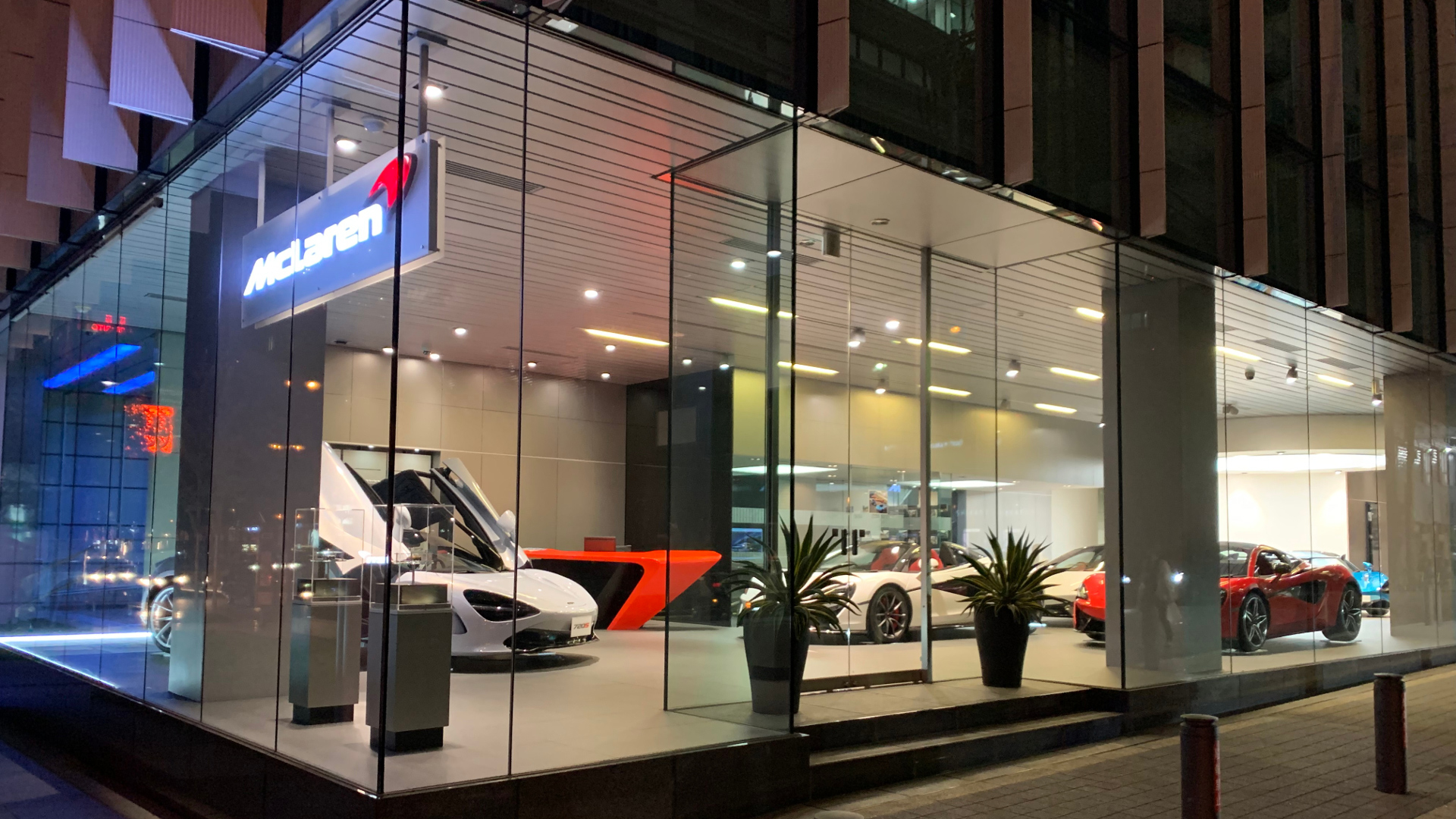 McLaren Osaka official retailer showroom exterior at night in Japan. A white McLaren 720S supercar is featured with its signature dihedral door open, alongside red and other luxury models inside the modern, glass-fronted dealership.