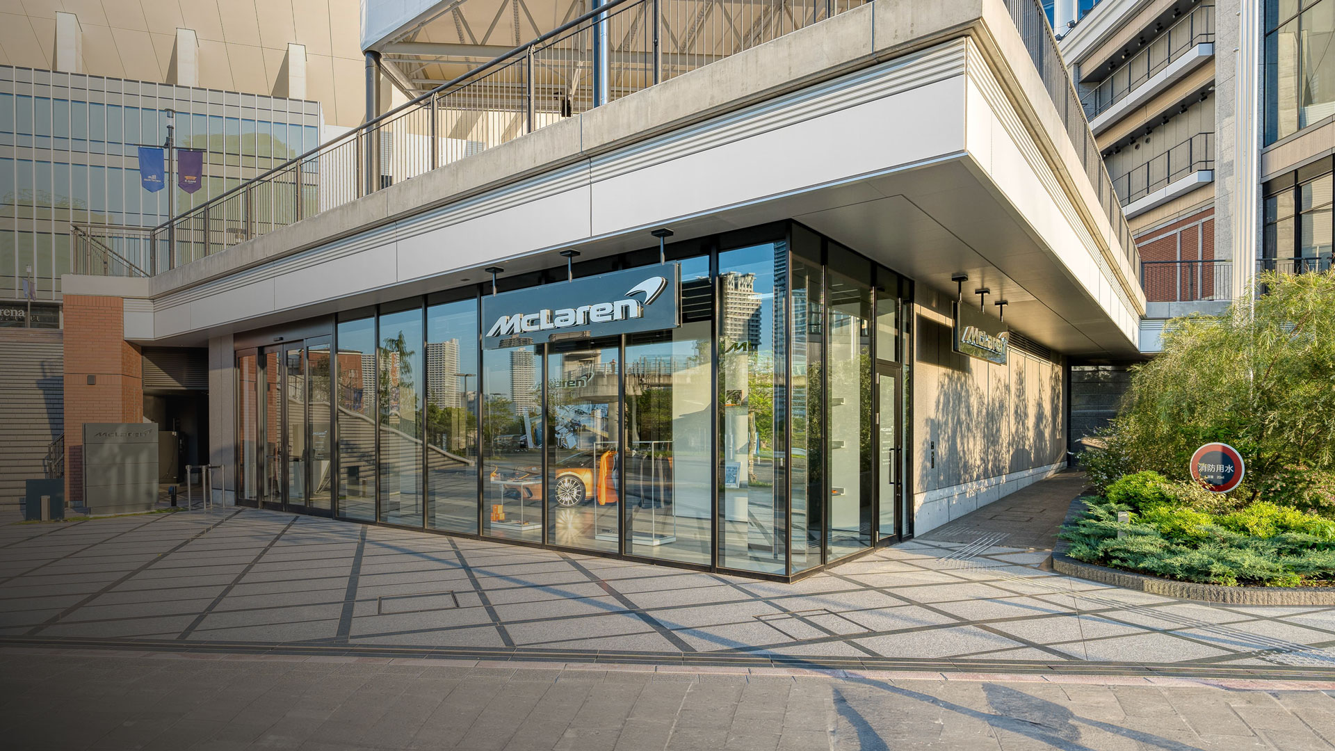 McLaren Yokohama official retailer exterior in Japan. The modern glass facade of the luxury car showroom is built into a commercial complex and displays a vibrant McLaren Orange supercar in the window.
