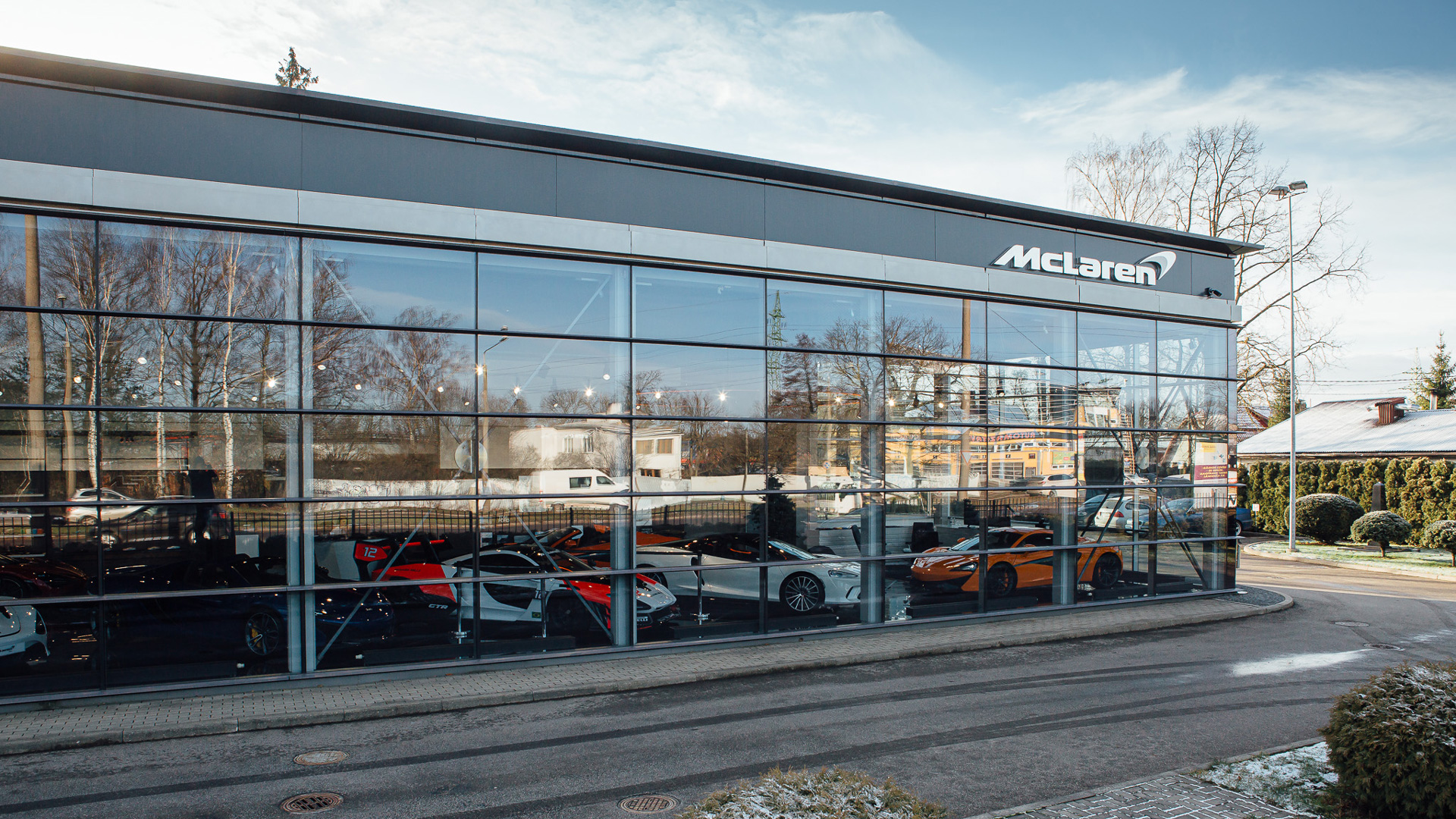 McLaren Riga official retailer exterior facade in Latvia, showcasing multiple supercars in the glass-fronted showroom. The inventory display includes white and orange McLaren 750S models and an orange Artura.