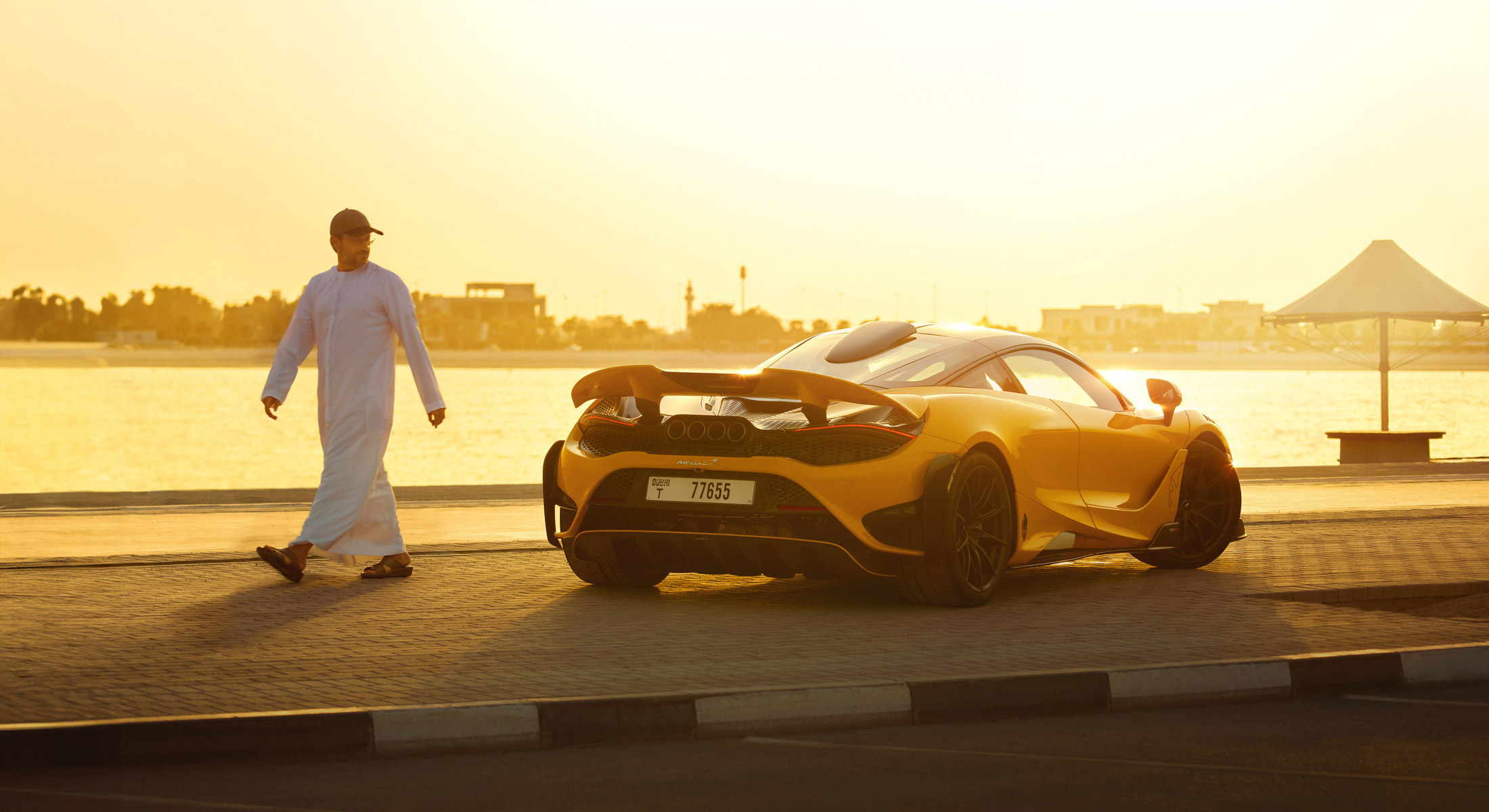 A striking rear-three-quarter view of a yellow McLaren 765LT parked by a calm body of water at sunset, with a man walking away from the car.