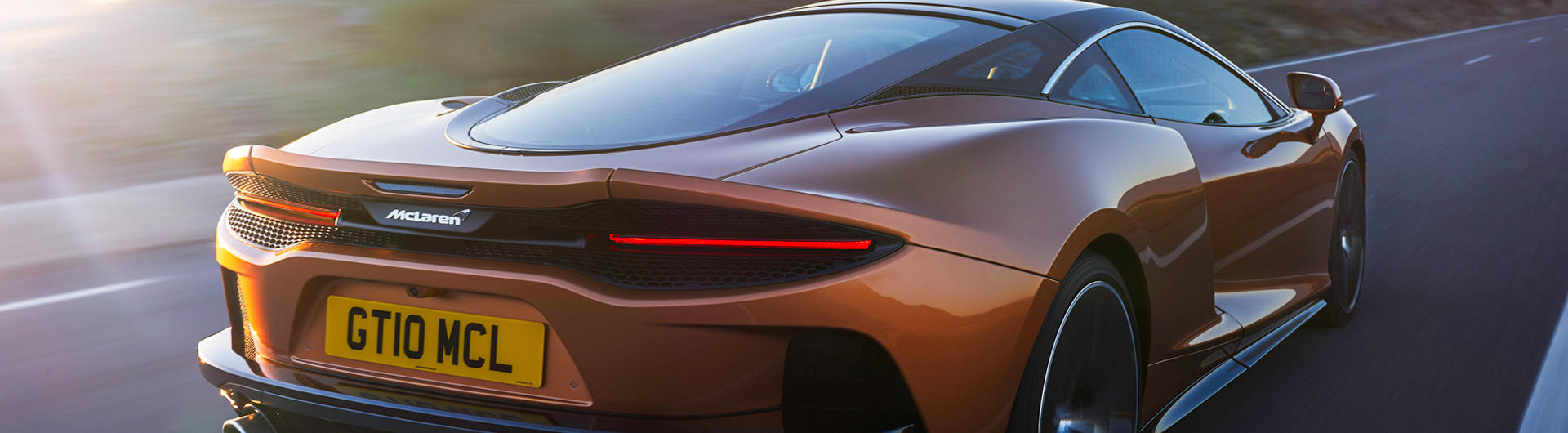 McLaren GT rear view on road, sunset orange paint, close-up, sports car driving, performance car.