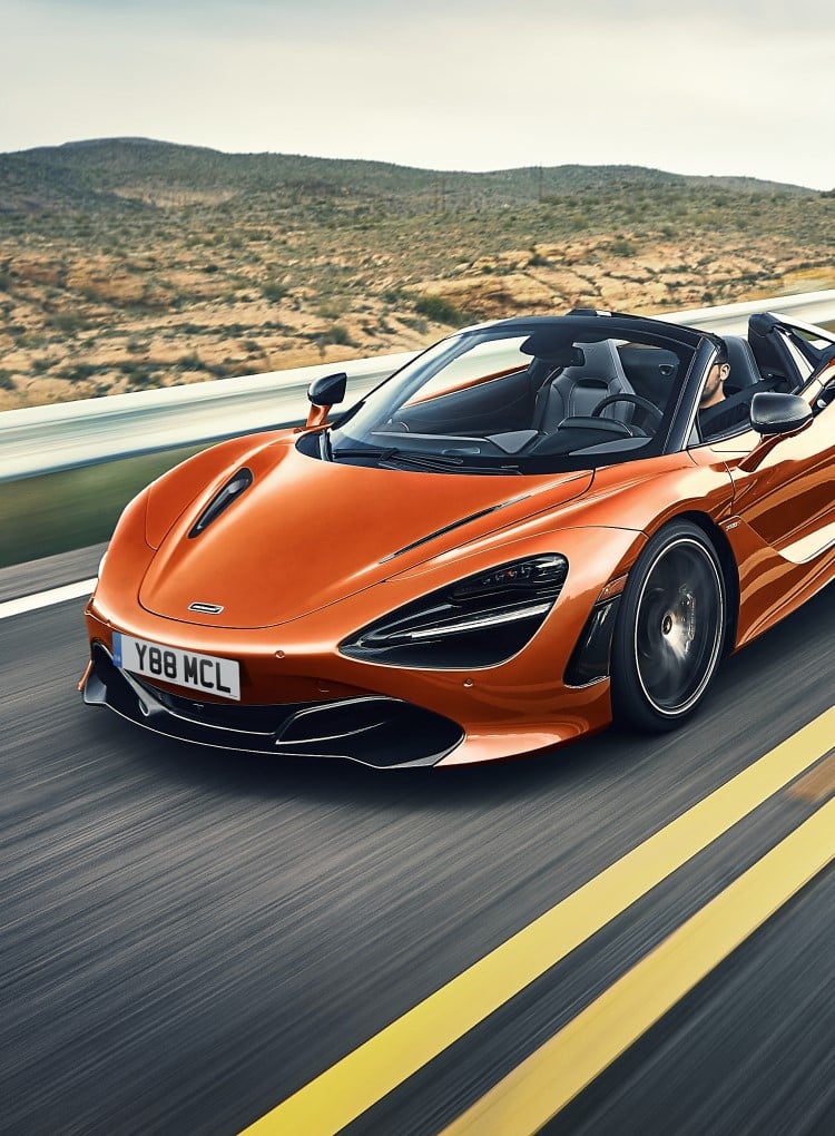 A front-side view of the McLaren 720S Spider convertible in a vibrant orange, driving on a road surrounded by a arid, rural landscape.