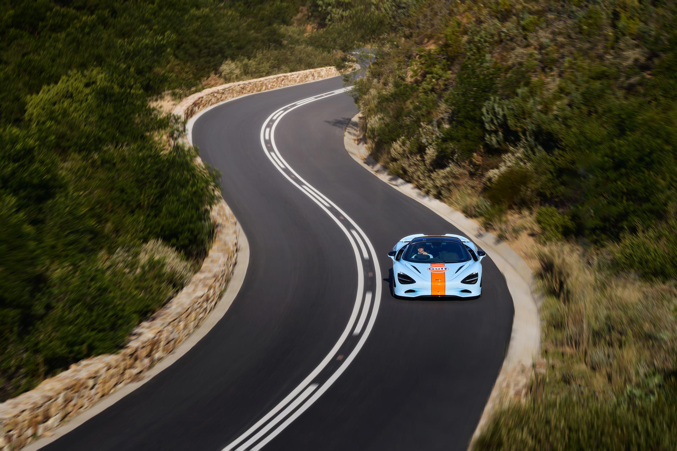 Front view Gulf Branded McLaren driving down a winding road, surrounded by trees.