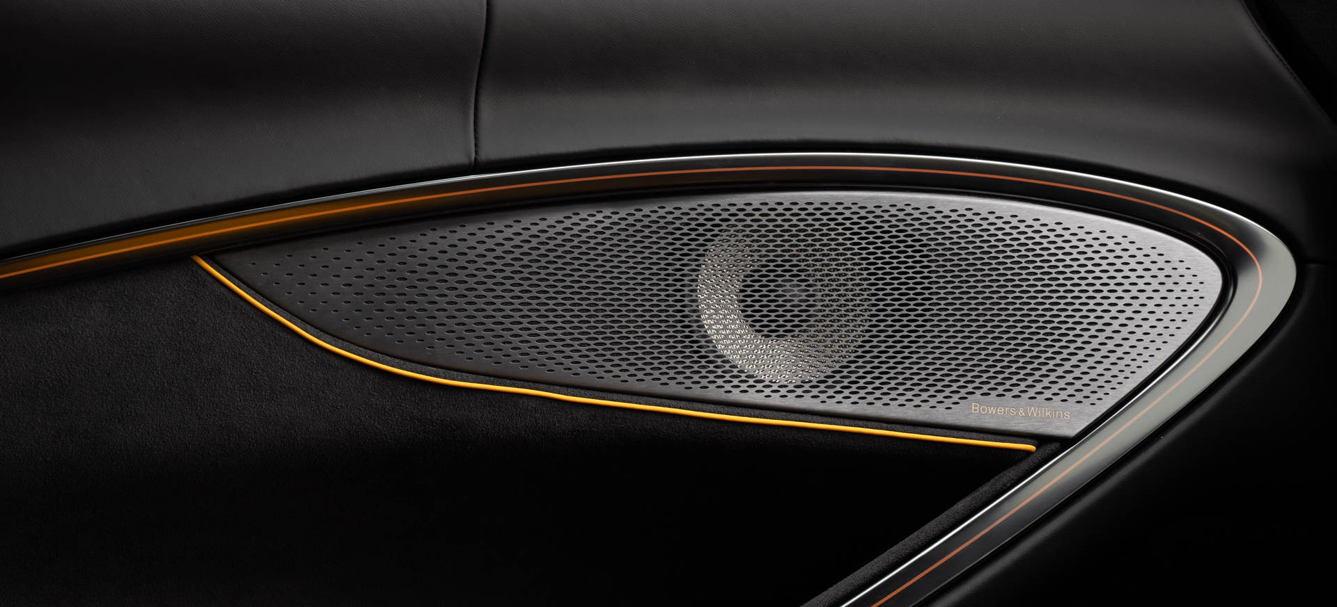Close-up of a premium Bowers & Wilkins audio speaker grille in a McLaren supercar door panel, featuring the distinct orange accent line, showcasing a technology partner collaboration.