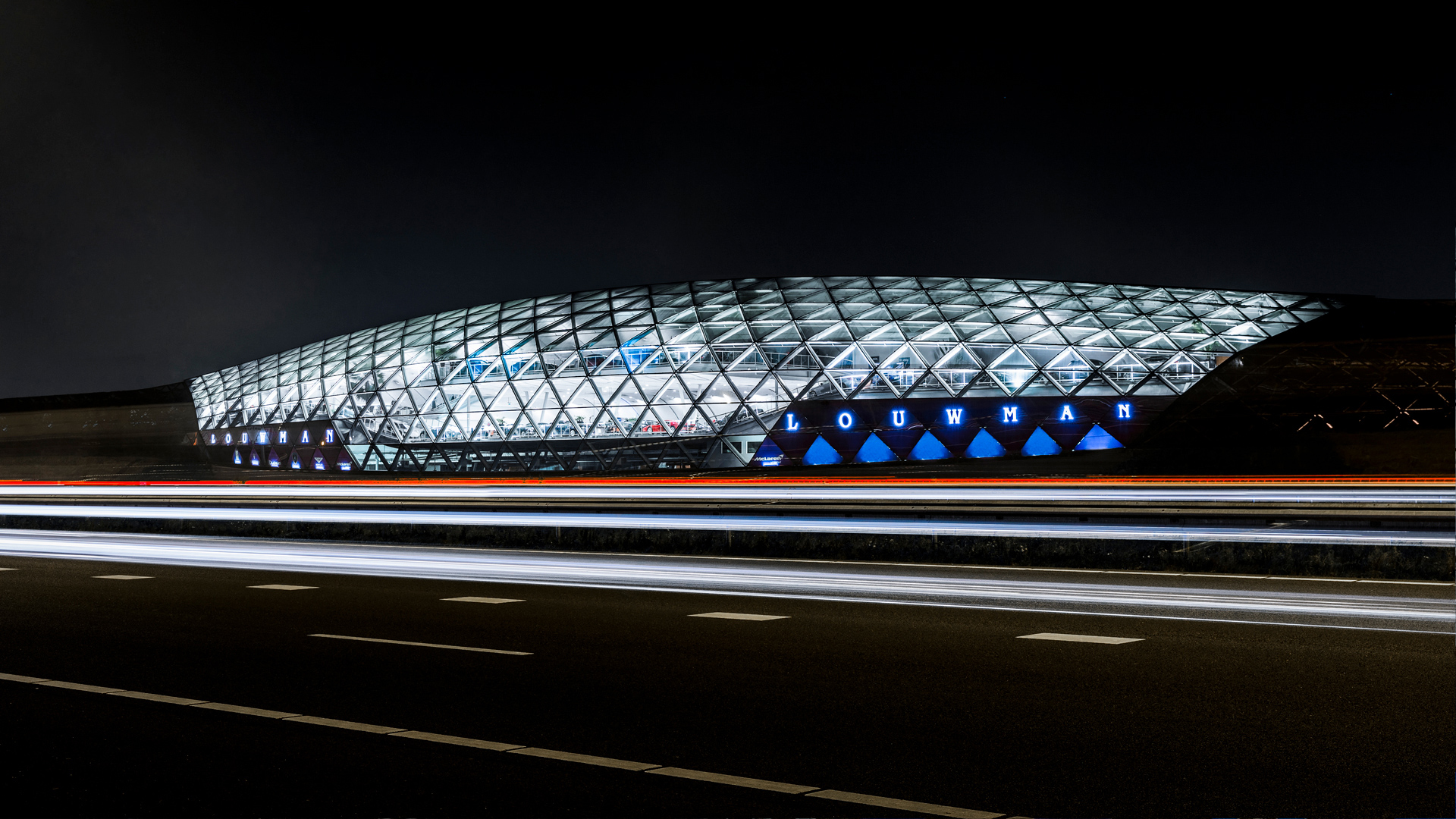 Long exposure night shot of the architecturally unique McLaren Utrecht dealership with striking illuminated glass facade and traffic light trails on the adjacent highway in the Netherlands.