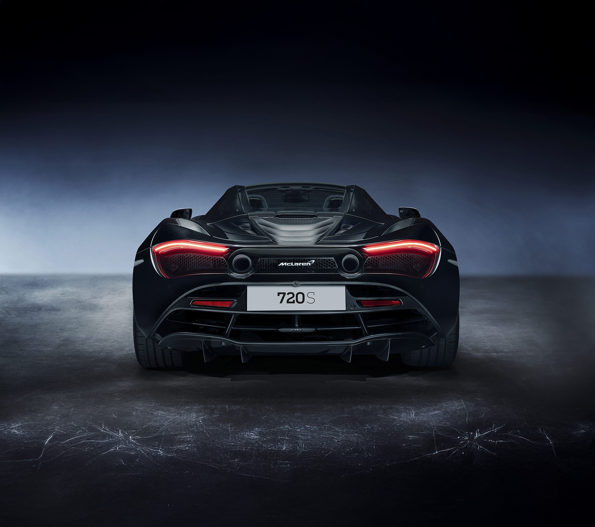 A rear view of a dark McLaren 720S Spider in a studio, highlighting its distinctive LED taillights, dual exhaust, and aggressive diffuser.