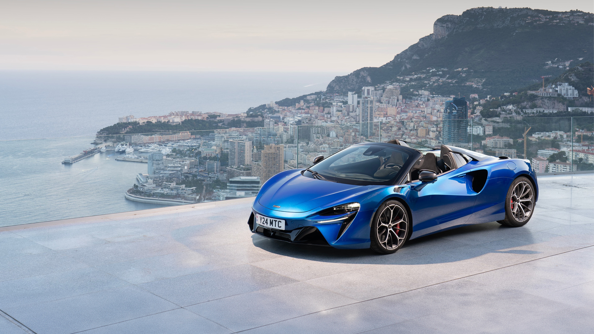 An atmospheric shot of the powerful blue McLaren Artura Spider parked on the apron of Monaco's yacht-filled port. The image contrasts the precision engineering of the plug-in hybrid with the luxury of the Riviera backdrop, setting the scene for pure performance.
