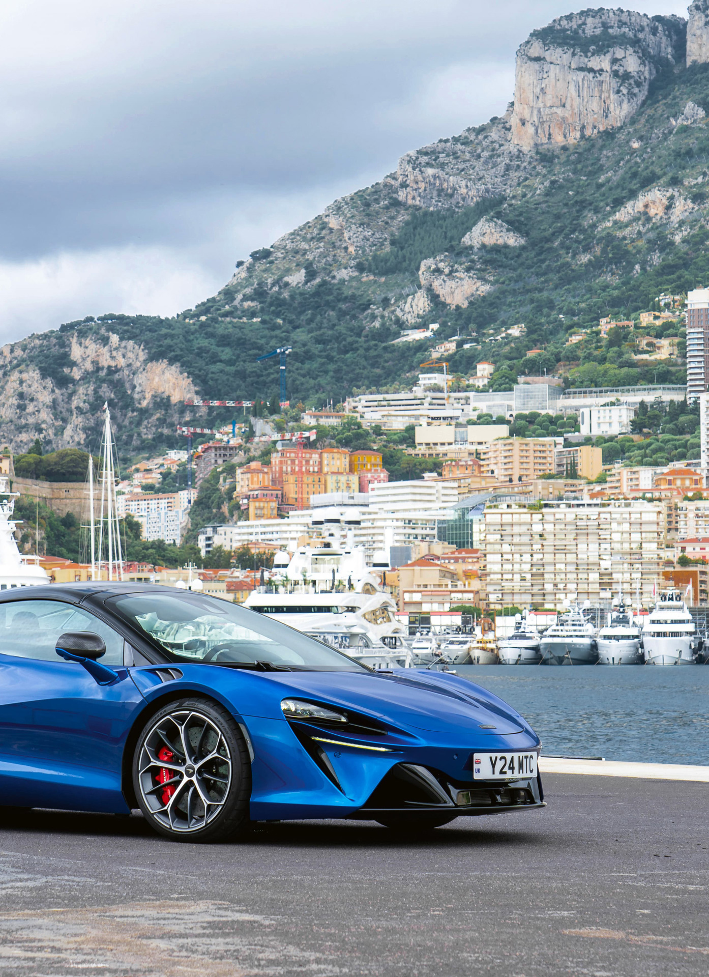 An atmospheric shot of the powerful blue McLaren Artura Spider parked on the apron of Monaco's yacht-filled port. The image contrasts the precision engineering of the plug-in hybrid with the luxury of the Riviera backdrop, setting the scene for pure performance.