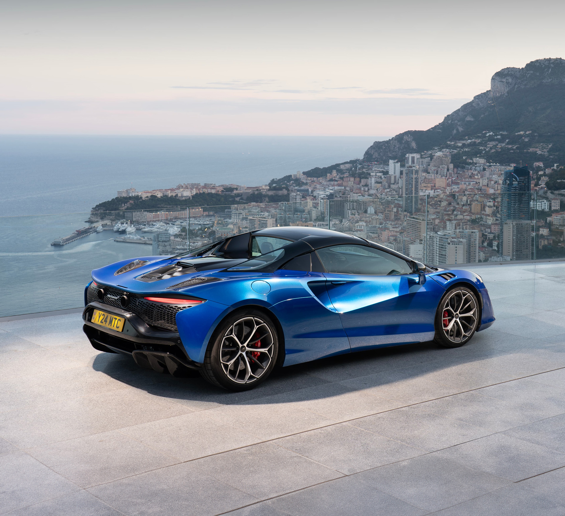 A stunning rear-quarter view of the Artura Spider hybrid supercar in Monaco Blue, perched on a terrace overlooking the city and the Mediterranean at sunset. This image highlights the car’s rear aerodynamics and elegant dihedral lines against the dramatic riviera coastline.