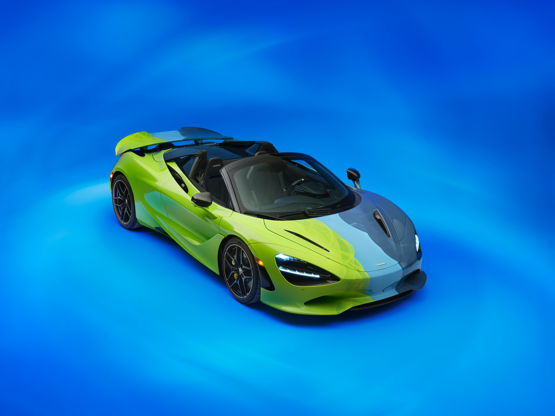 The Project Chromology 750s by MSO and Nat Bowen, on display against a glowing blue background. The car is various overlaying shades of green and blue and can be seen parked at an angle with its headlights on.