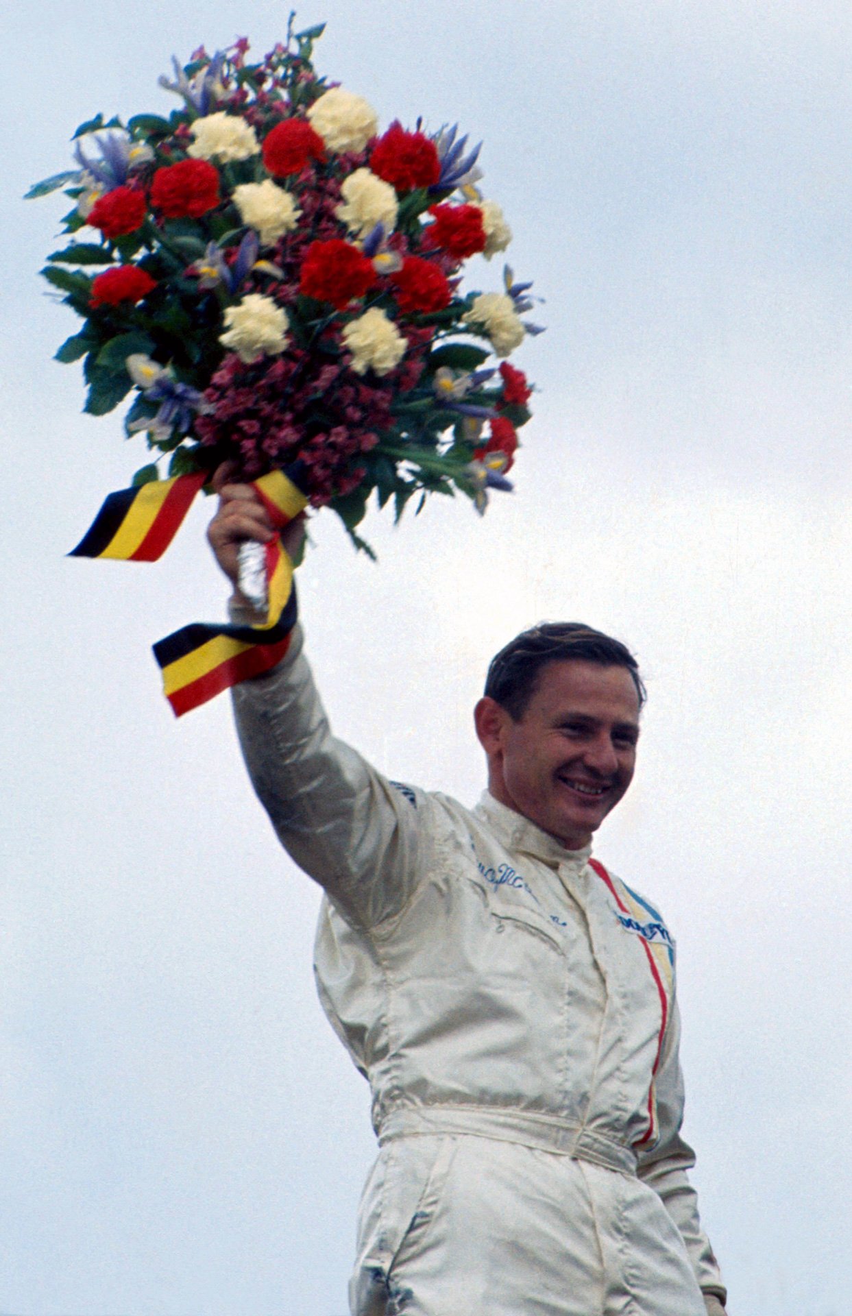 Bruce McLaren holding up a bouquet of flowers whilst wearing his racing suit.