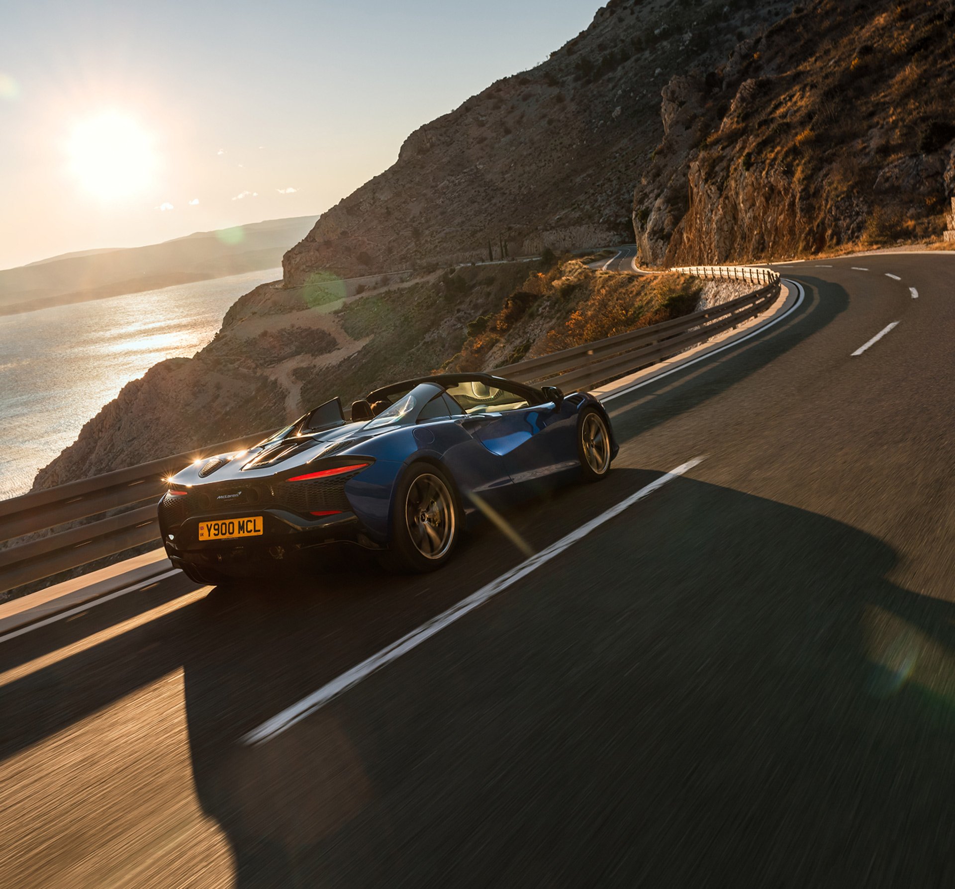A synamic shot of the McLaren Artura Spider driving at speed along a road with the sun setting in the background.