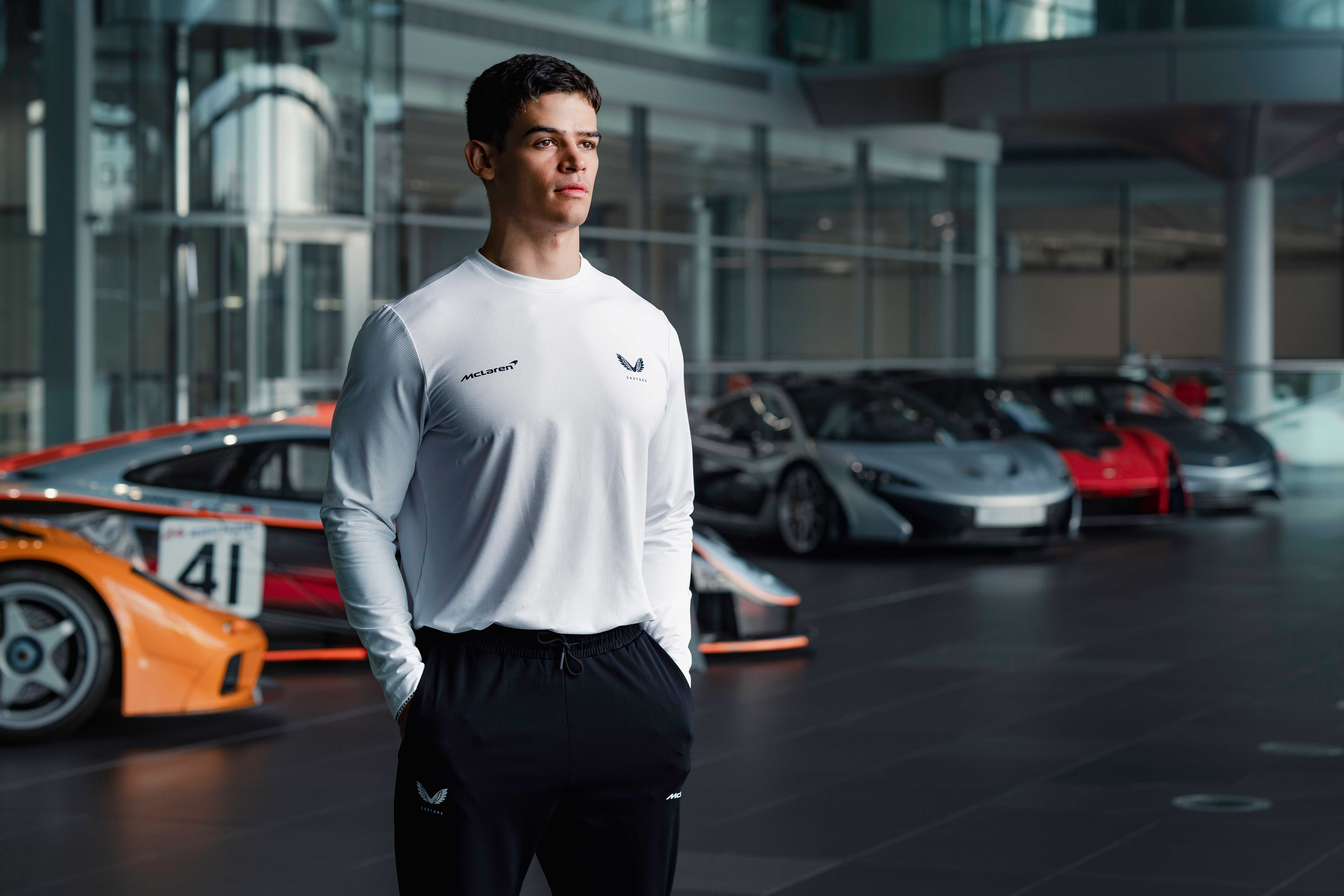 Male model wearing a white long sleeve McLaren Castore top and black McLaren Castore joggers, stood in the McLaren technology centre in front of the cars.