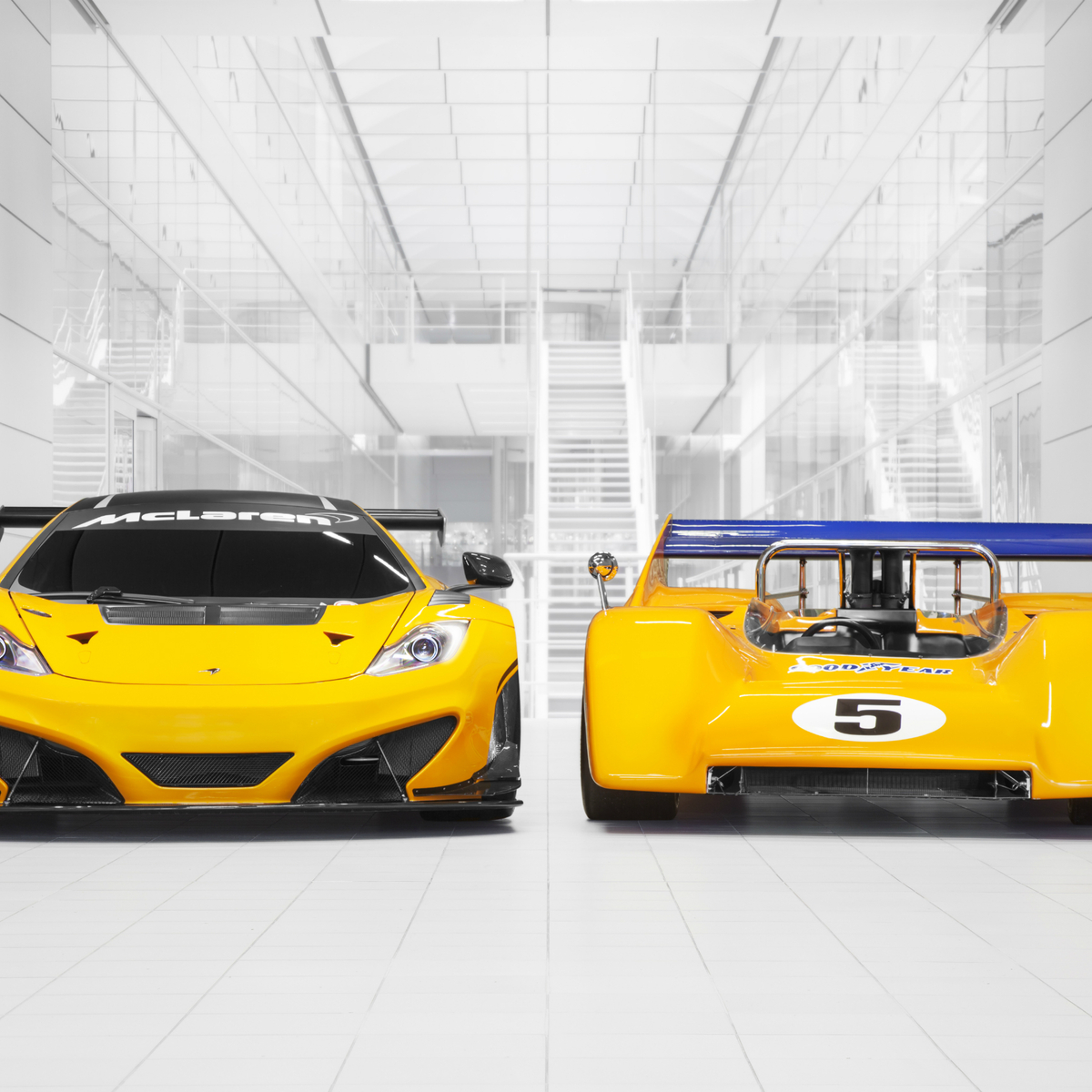 Two bright yellow McLaren 12C GT Can-Am Edition, one facing forward and the other facing away, in a clean, white building. 