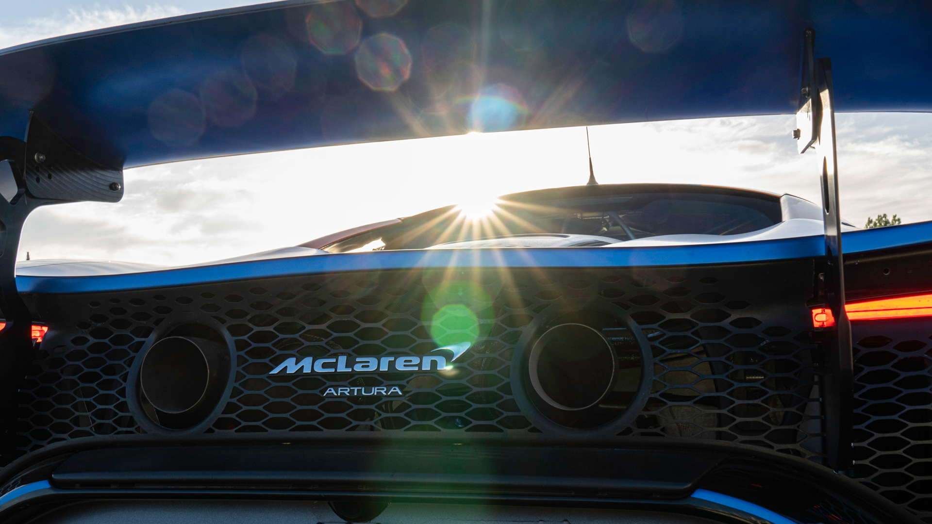 Rear close up of a McLaren Artura GT4 parked on a track, with the sun behind it.