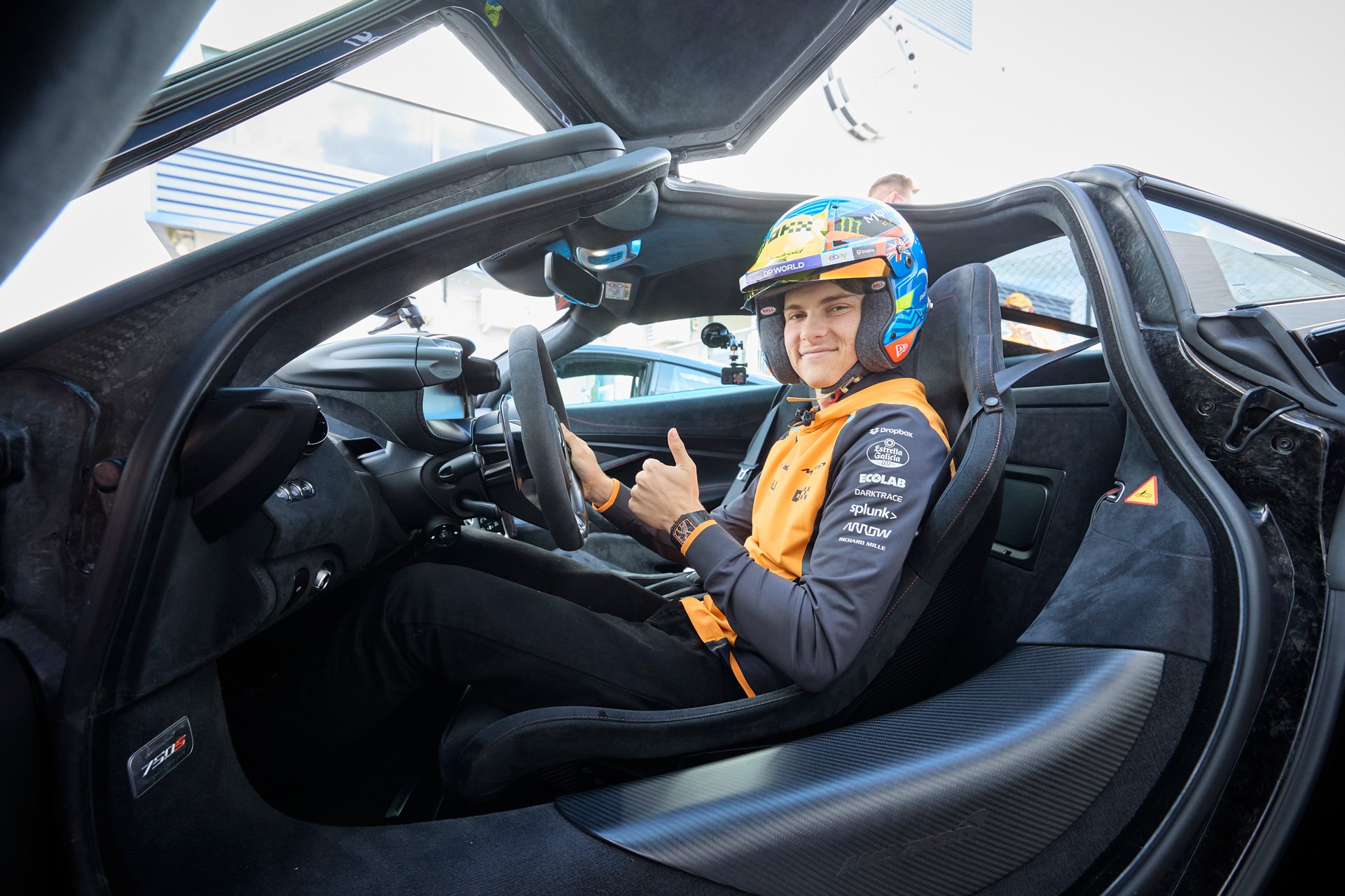 Oscar Piastri sat in the drivers seat of a McLaren 750S, wearing a helmet, and signalling thumbs up.  