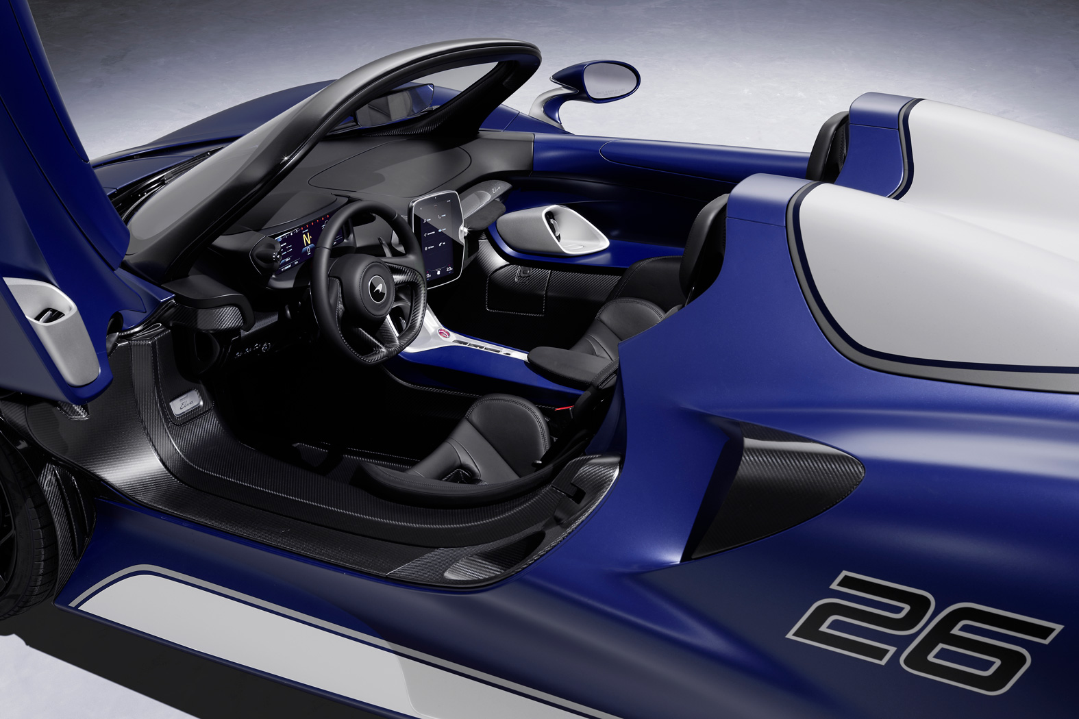  A view of the interior of a blue McLaren Elva, highlighting the black leather seats, digital infotainment screen, and carbon fiber trim.