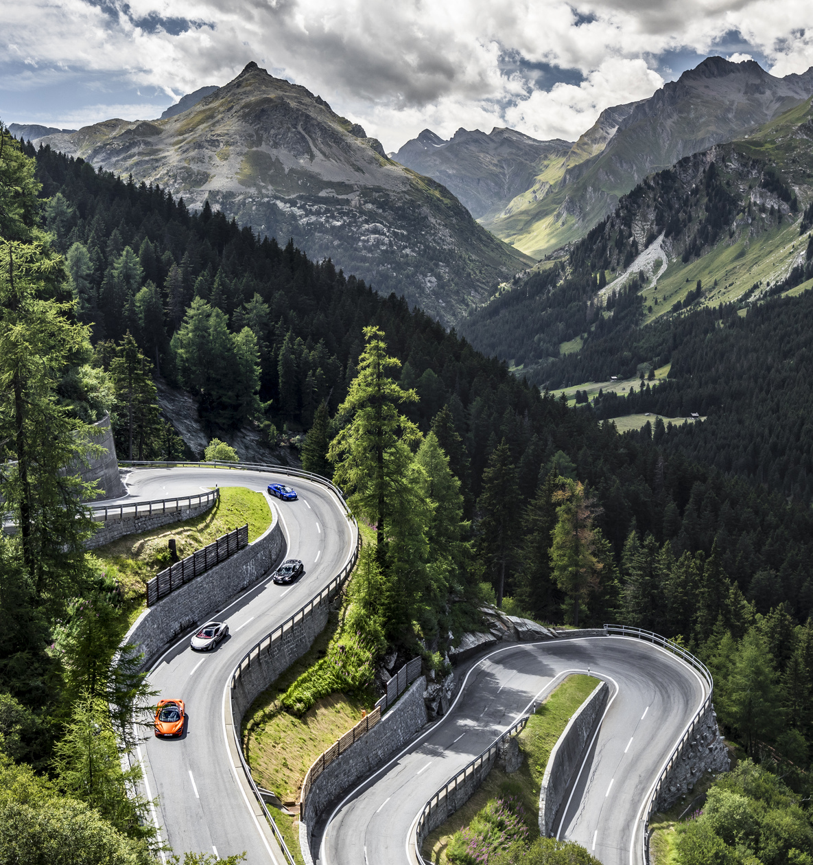 An aerial view of a winding mountain road with four McLaren supercars (orange, grey, black and blue) navigating hairpin turns amidst a lush green forest and towering peaks under a cloudy sky.