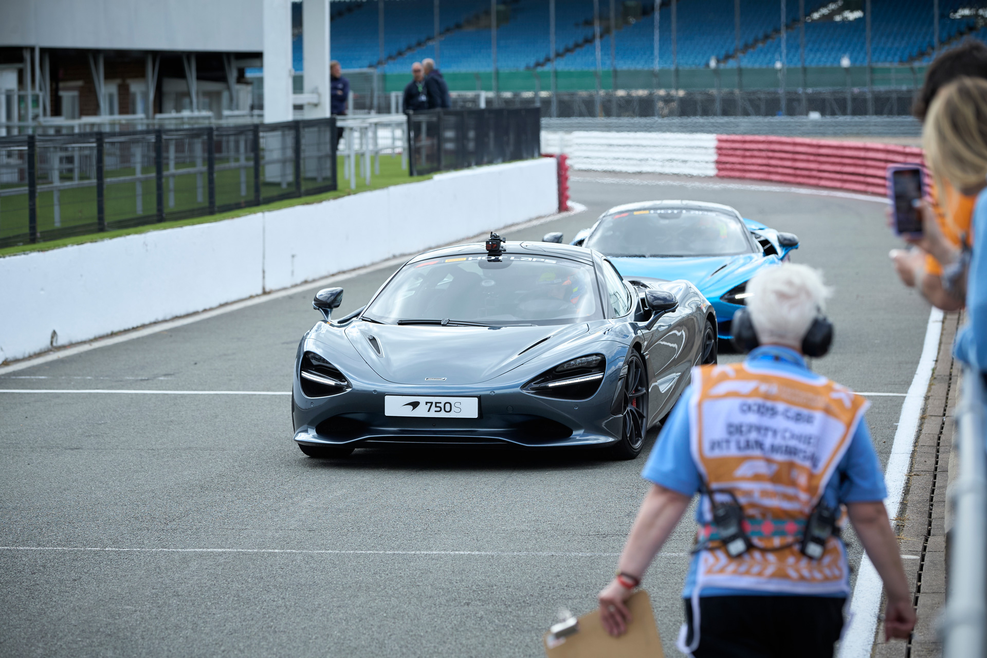 Silver McLaren 750S supercar on a racetrack, with a race marshal in the foreground.