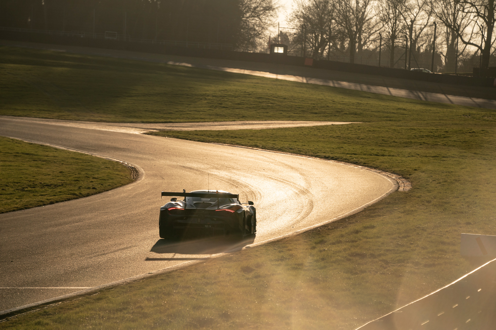 Distant shot of a McLaren 720S GT3 Evo driving away around a winding race track at sunset.
