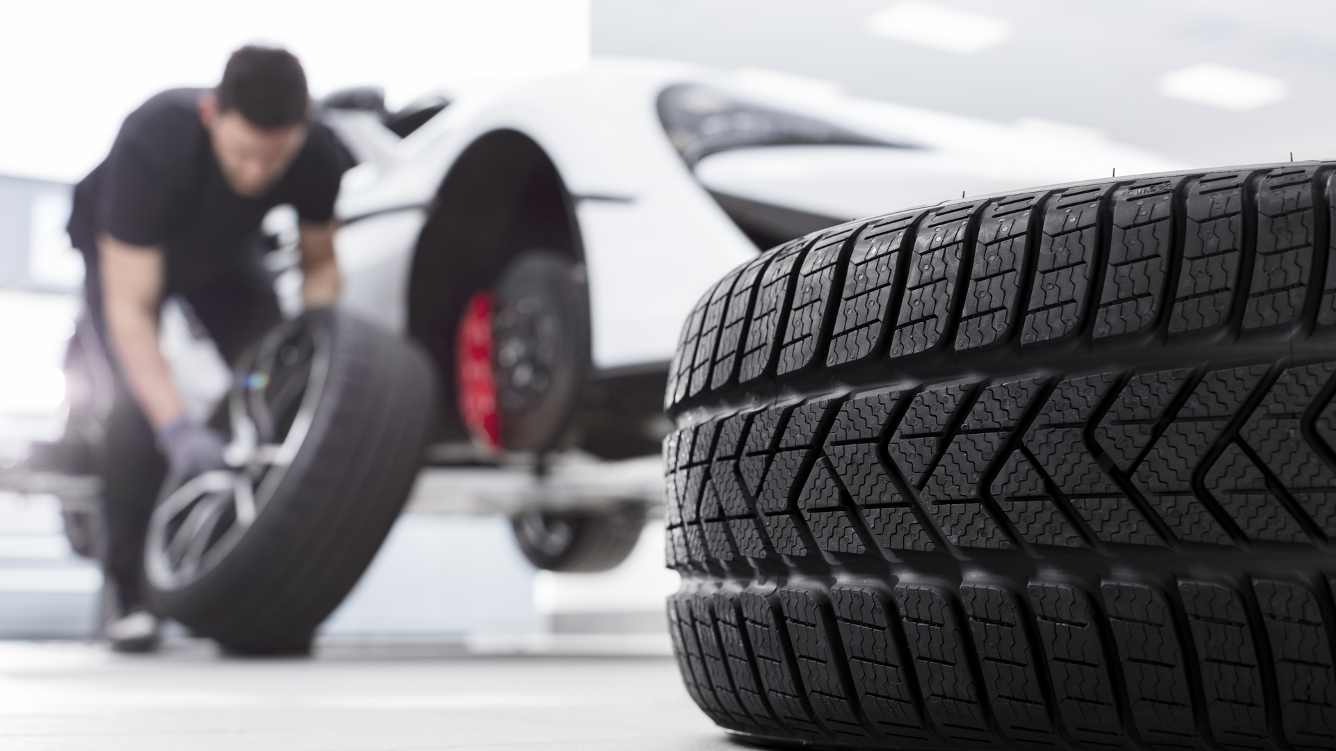  A close-up of a new tire in the foreground, with a mechanic in the blurred background working on a white McLaren car with a red brake caliper.