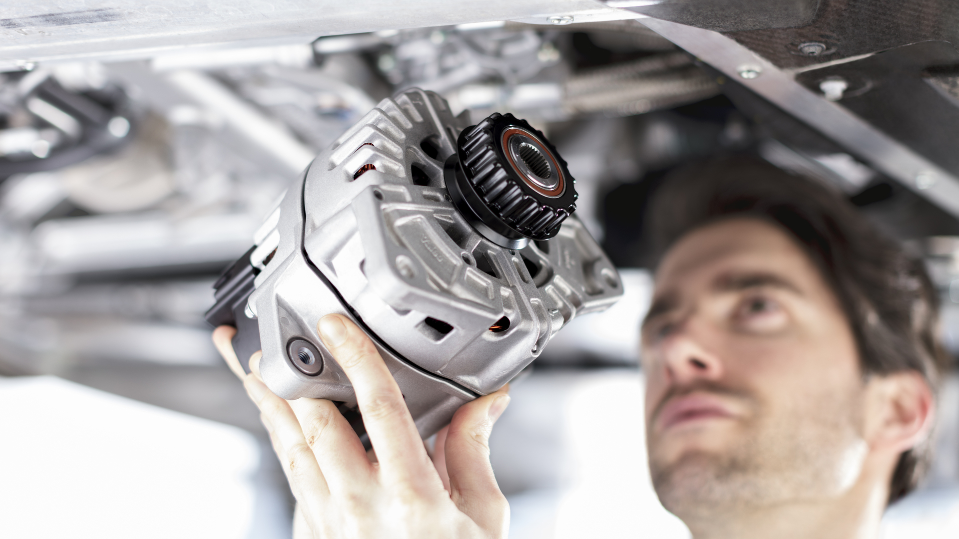 A male McLaren engineer holding and inspecting an alternator while working on a car.