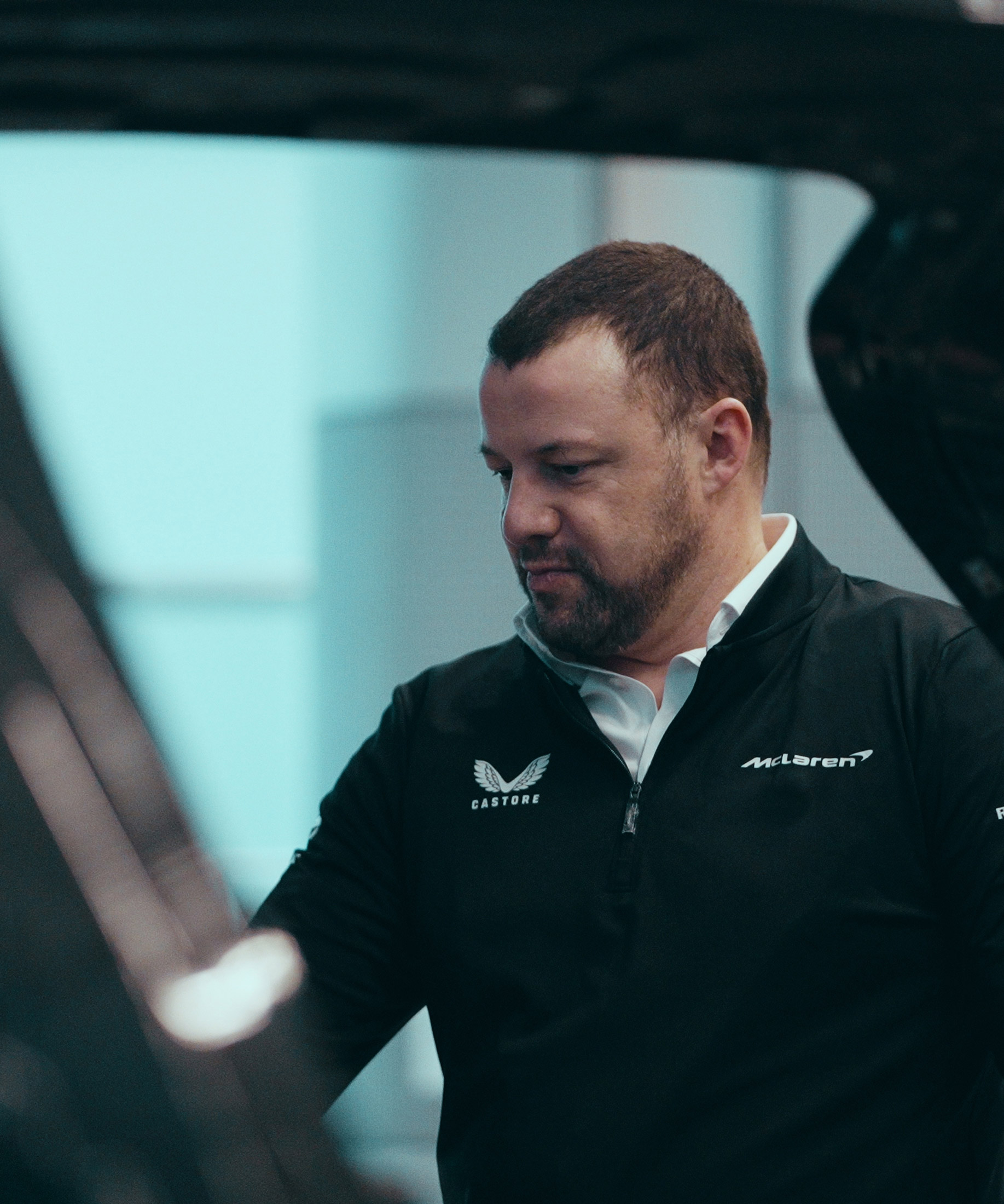 A man with short hair and a beard, wearing a black McLaren jacket, looks downwards while standing next to a car, with a blurred light background.