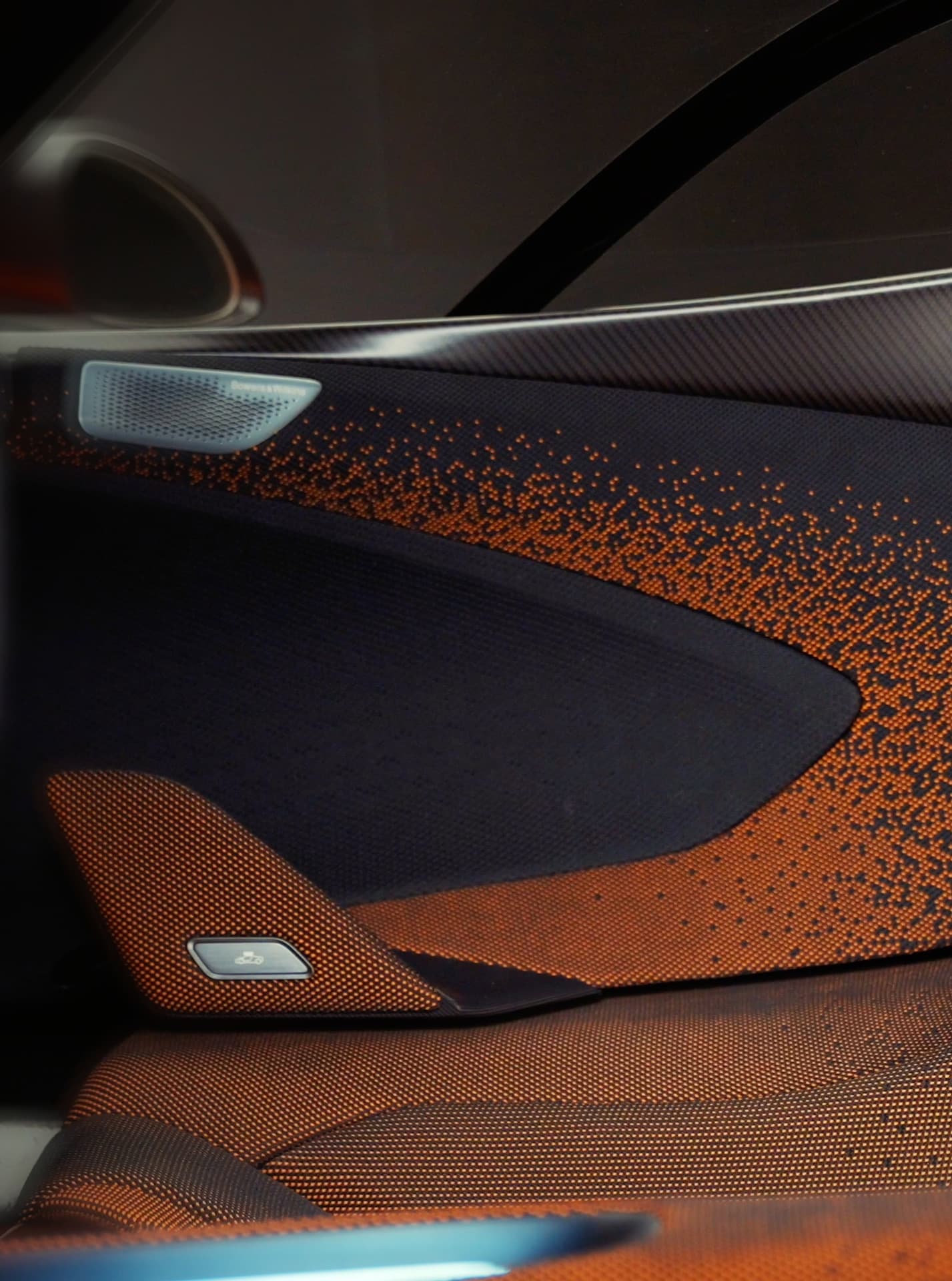 A close-up vertical shot of a McLaren car's interior door panel, featuring a unique black and orange pixelated fabric design, carbon fiber trim, and a small speaker grille.