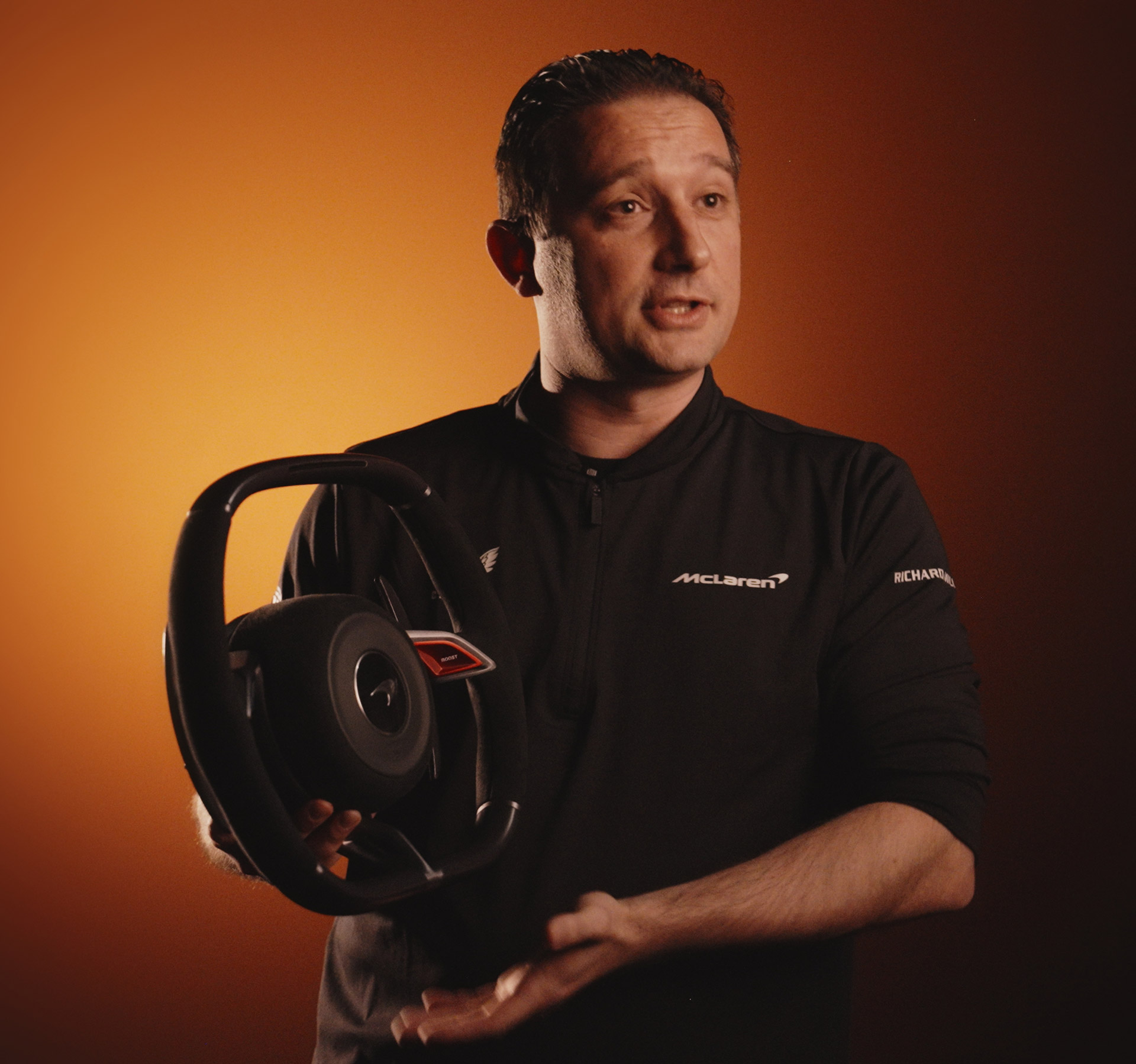 A man in a black McLaren-branded jacket presents a car steering wheel, looking towards the viewer and speaking.