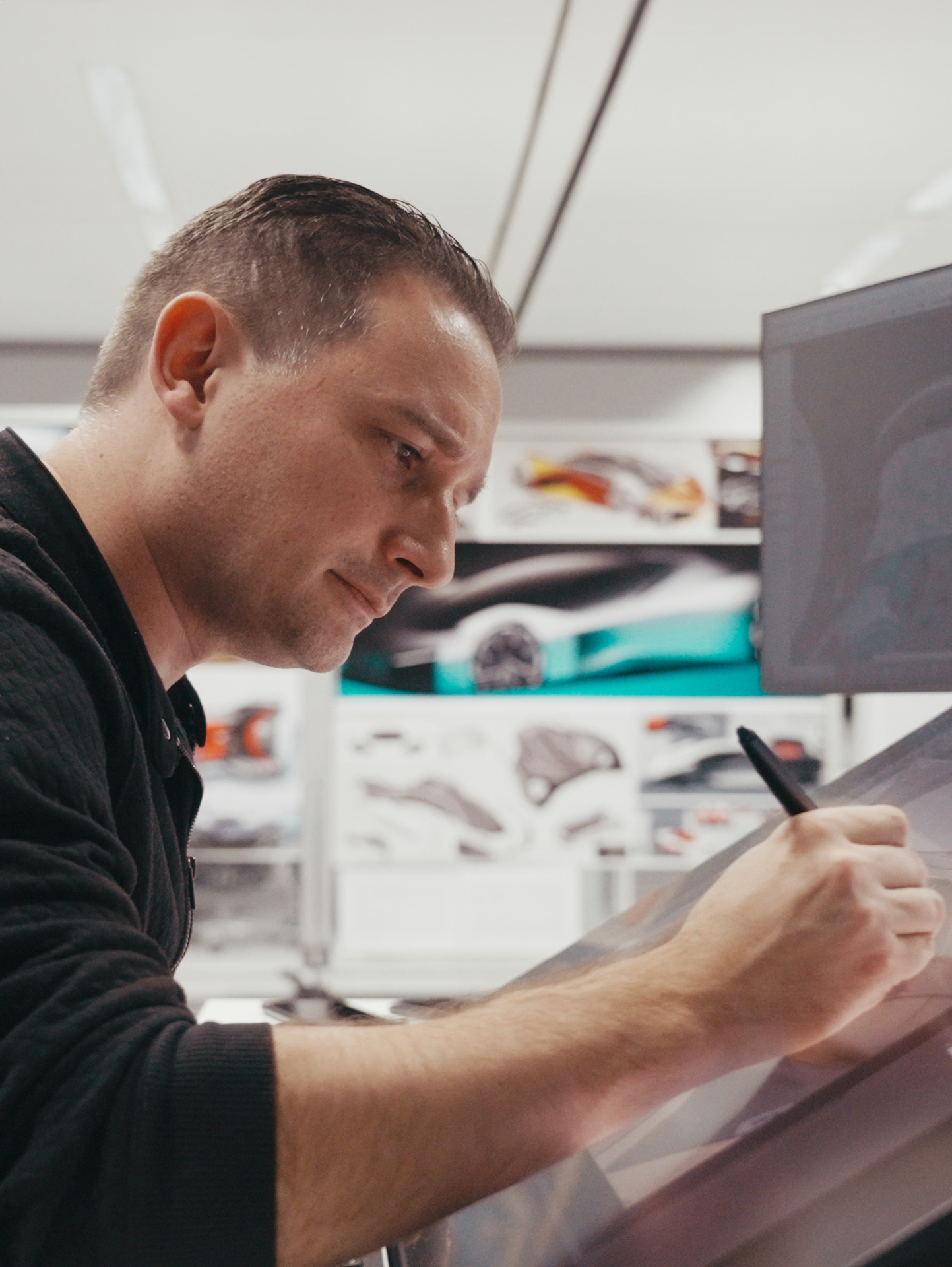 A man with short hair is intently sketching on a digital tablet, with car design concepts visible on a screen and wall in the background.
