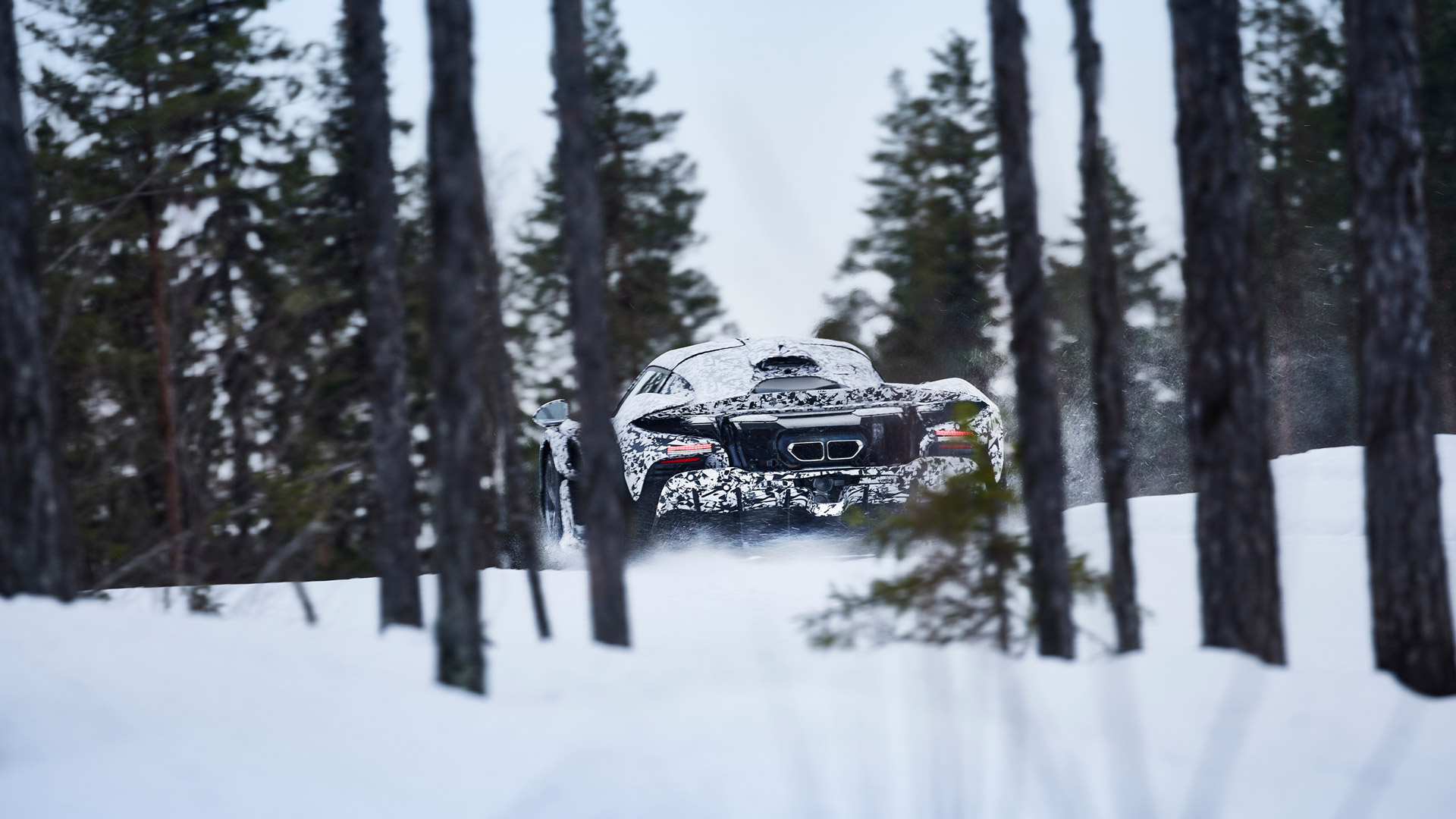A camouflage-wrapped McLaren W1 prototype drives away on a snowy path, seen between a dense line of pine trees.