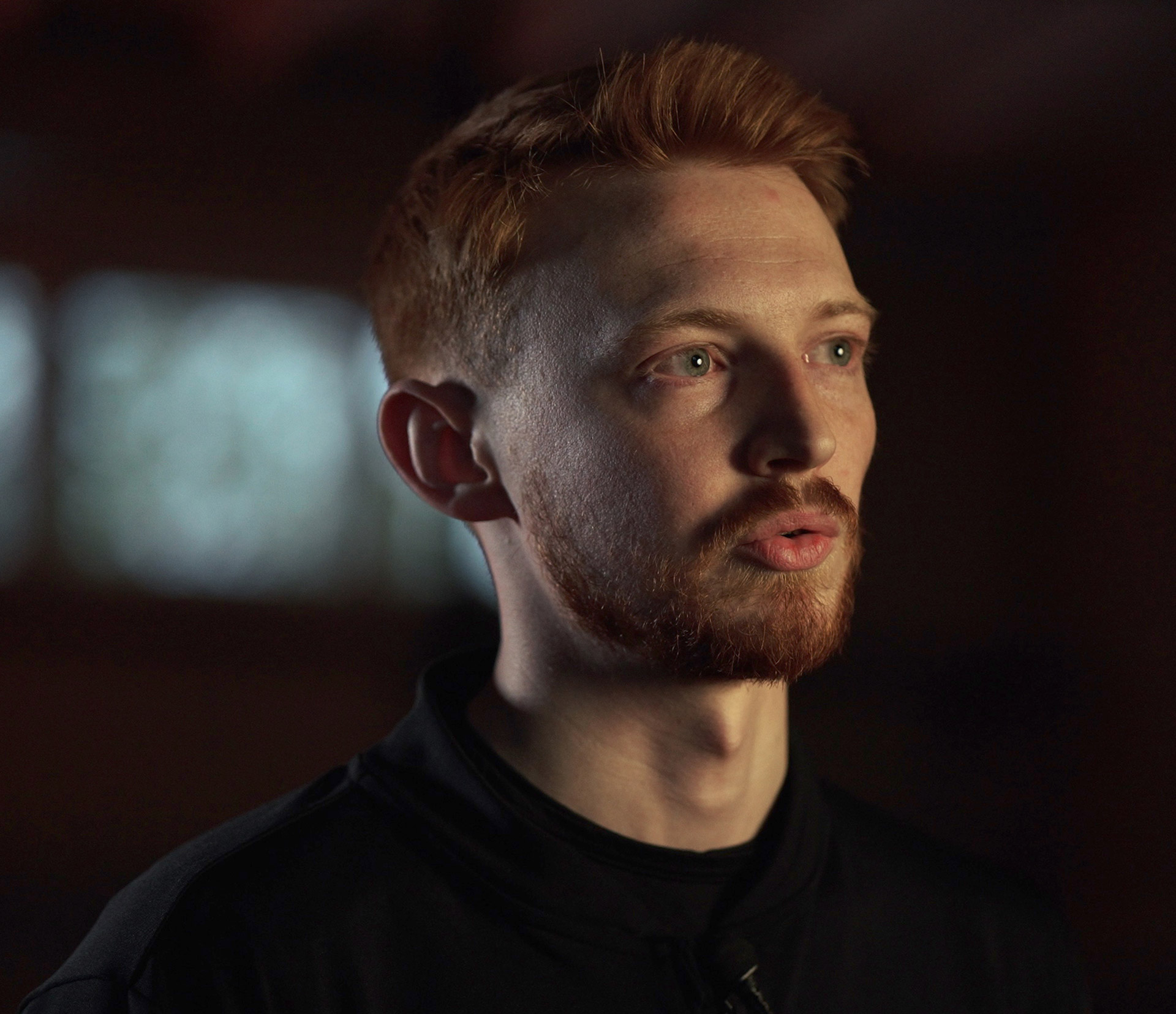 A close-up portrait of a person with red hair and a beard, looking slightly upwards and to the right, with a dark background.