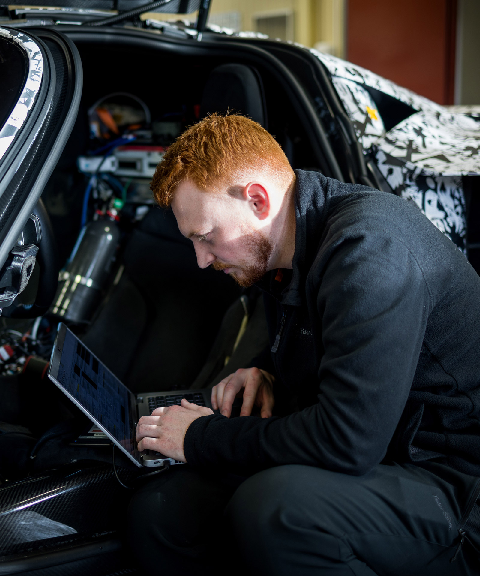 A person with red hair and a beard sits in the open door of a camouflage-wrapped car, focused on a laptop.