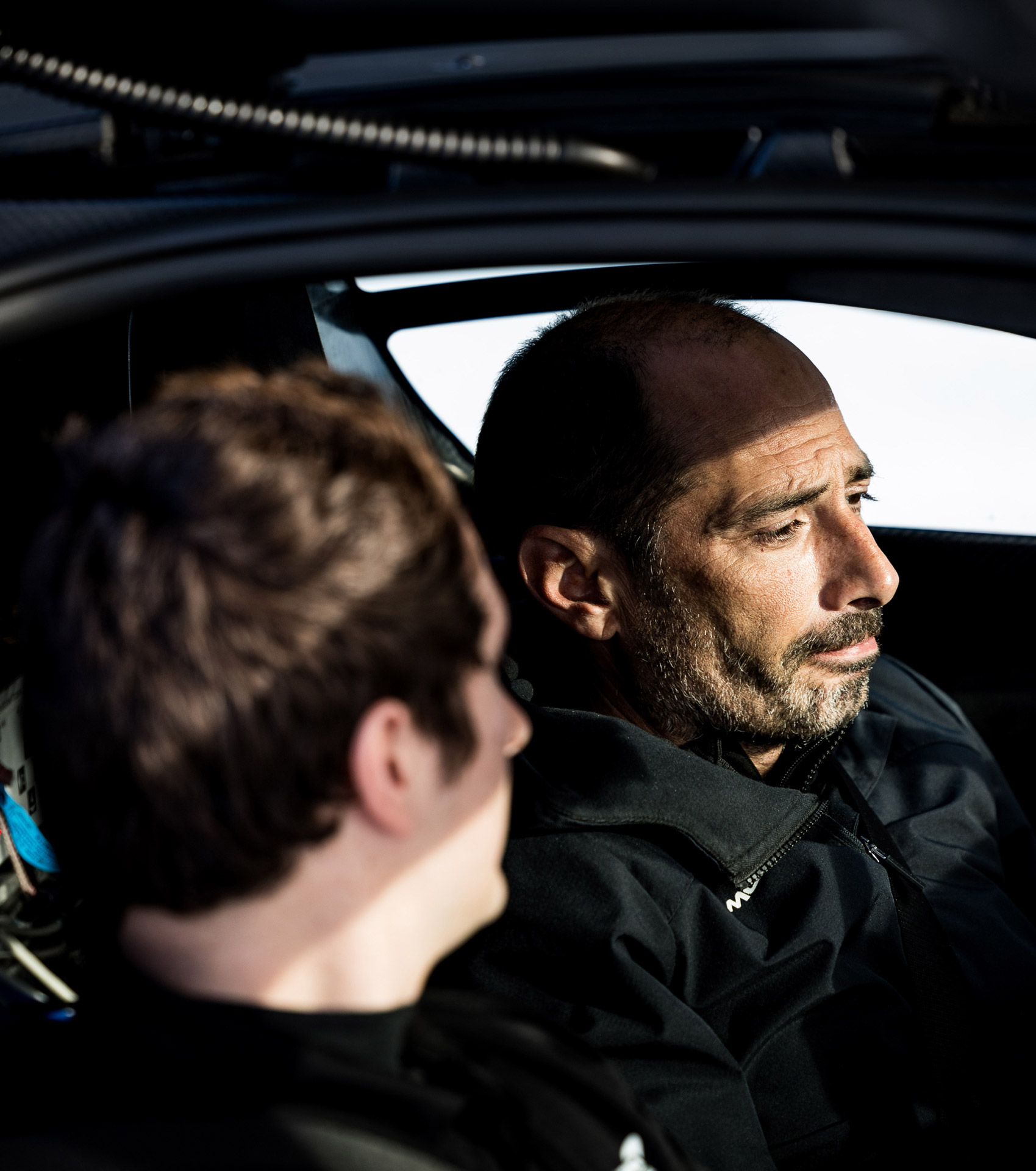 A side profile shot inside a car showing two men, one with a beard looking intently out the window, and another with the back of his head visible.