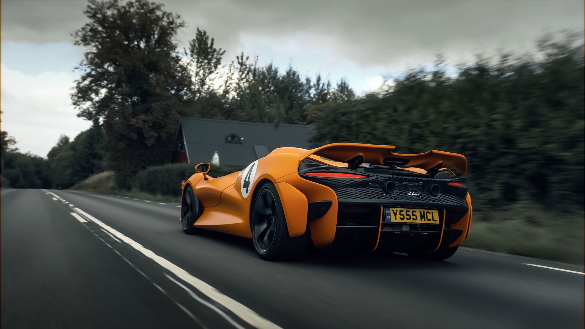 An orange McLaren Elva with a racing number, driving down a rural road.