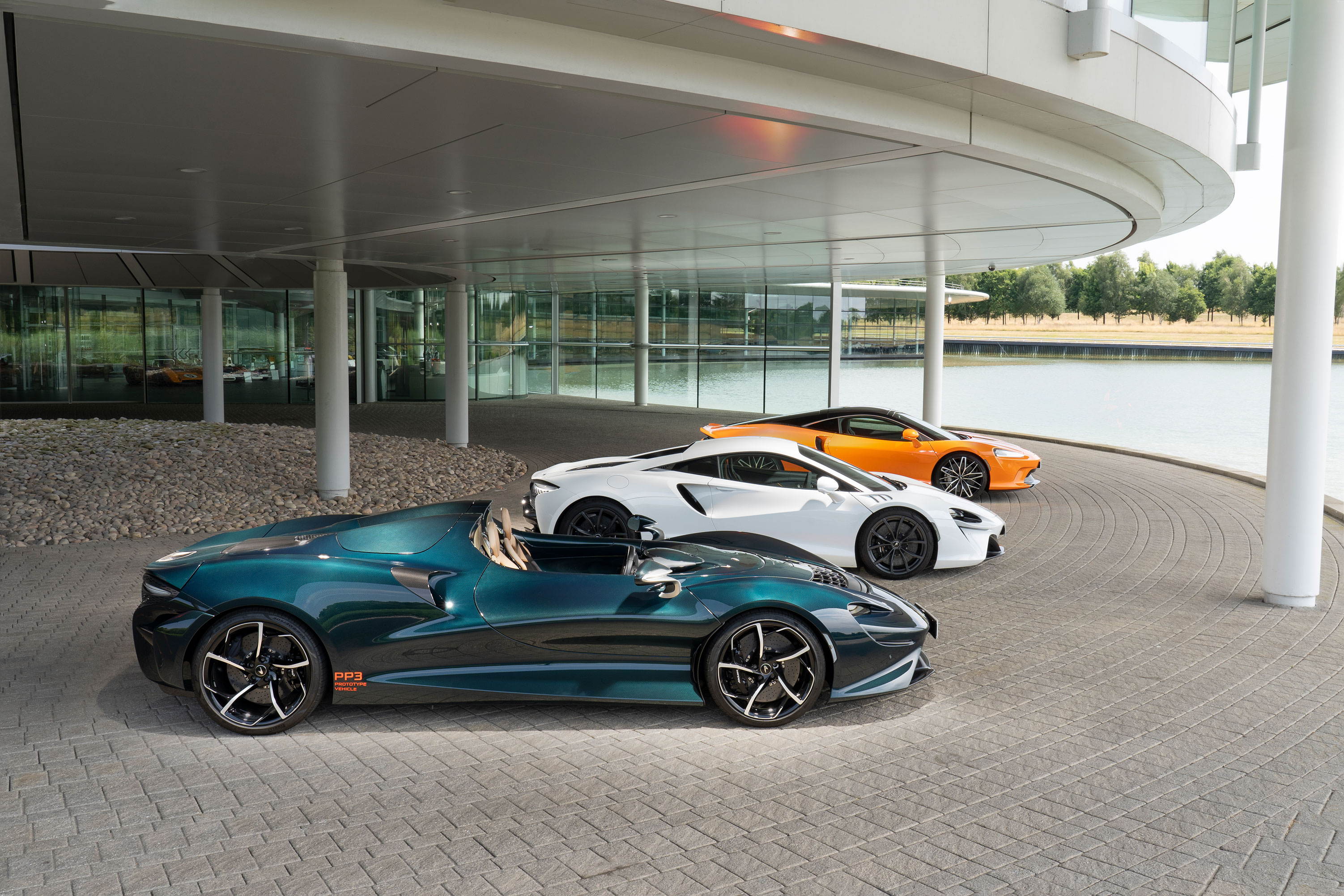 Three McLaren supercars, including a teal convertible, white coupe, and orange coupe, parked outside the McLaren Technology Centre.